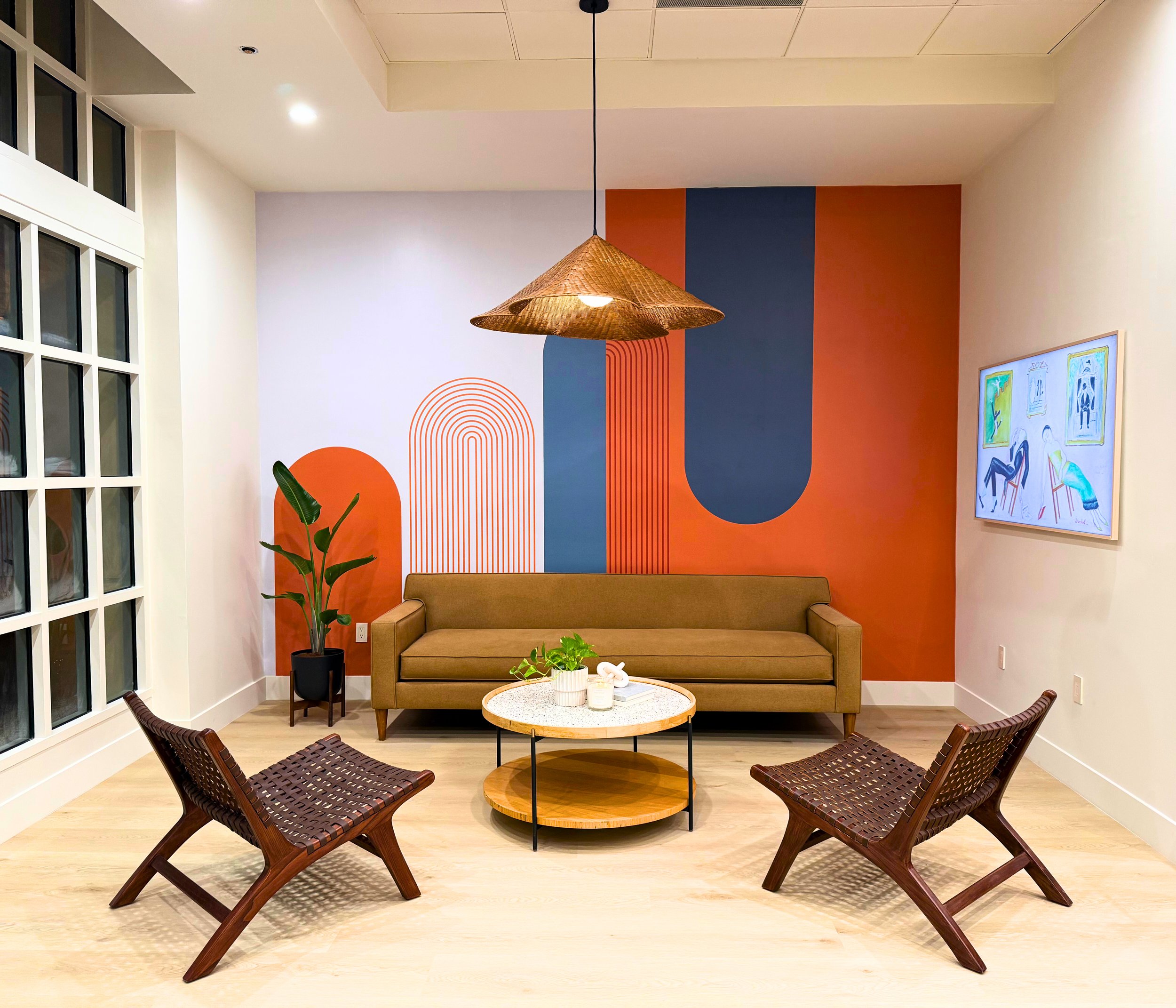 A modern living room with a tan sofa, two wooden lounge chairs, and a round marble coffee table. The back wall features geometric art in shades of orange, blue, and white. A large window on the left and a framed art piece on the right wall add to the