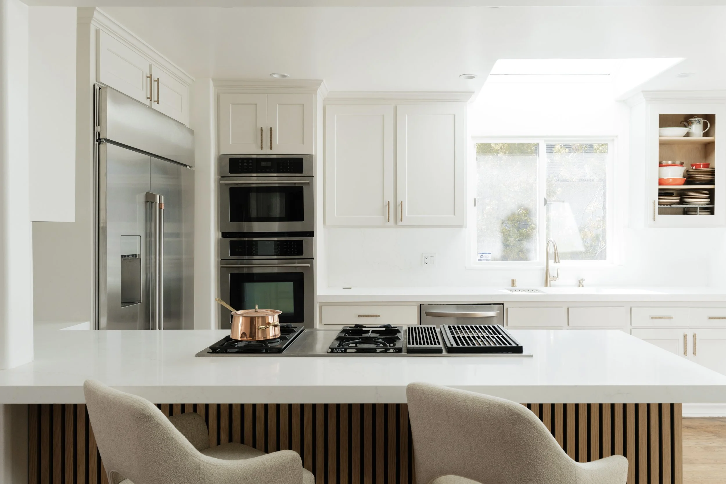 Refinished modern white kitchen with stainless steel appliances, including a double wall oven and refrigerator, a white countertop island with a copper pot, open wall cabinets with dishes, a window above the sink, and a light-filled space.