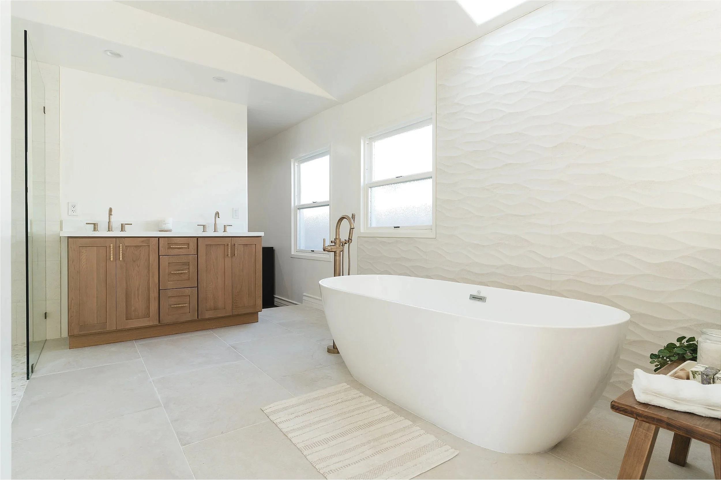 Modern bathroom remodel with a freestanding white bathtub, custom wooden vanity with dual sinks, minimalist decor, textured white wall, and neutral-colored tiles.