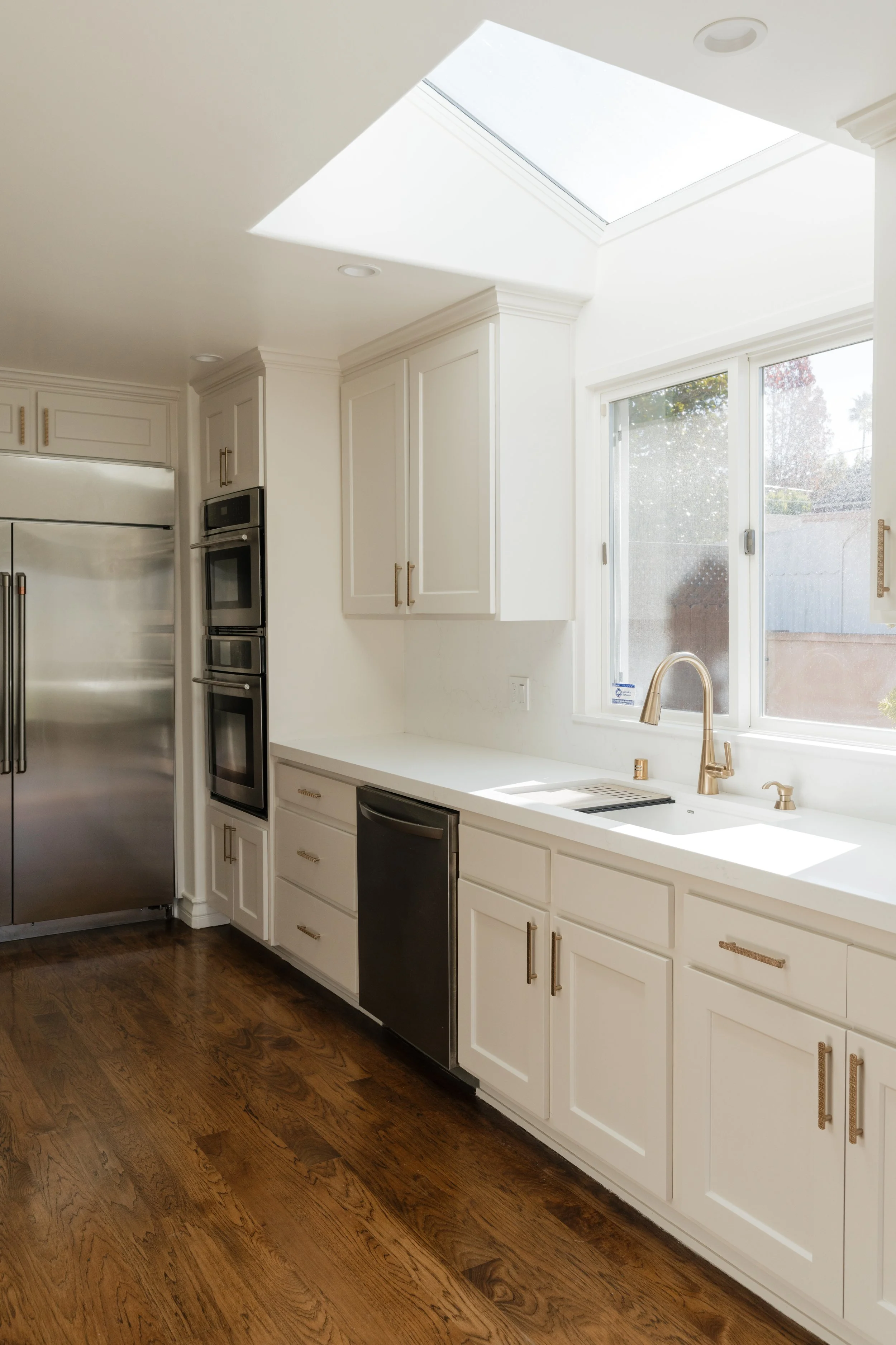 Bright kitchen with white cabinets, stainless steel refrigerator, double oven, black dishwasher, and a large window with a view of the outdoors. Wooden floor and a skylight above the sink.