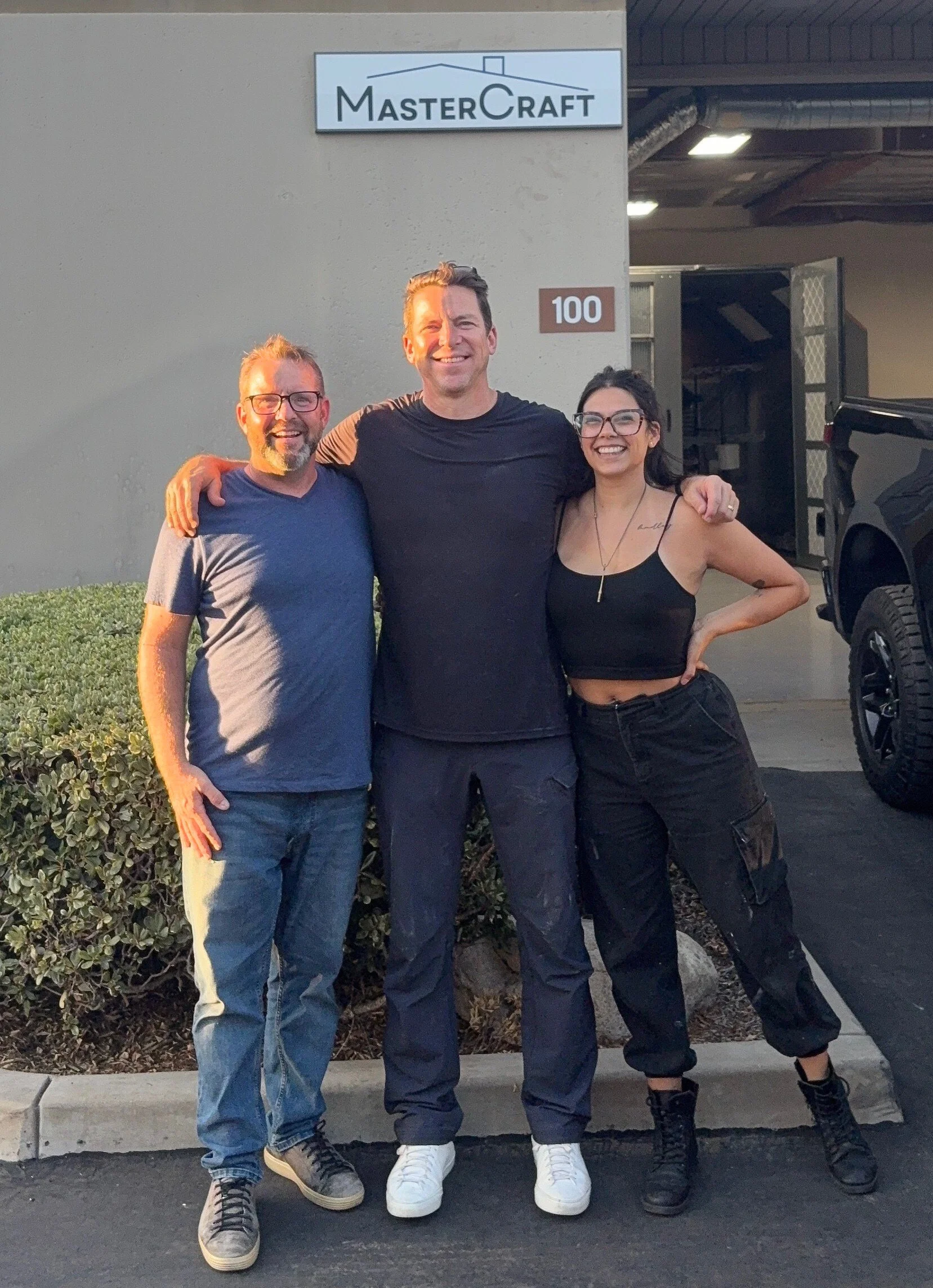 Three smiling contractors standing together outside a shop with a sign that reads 'MasterCraft,' during the evening. The person in the middle has his arms around the other two, and they are all looking at the camera.