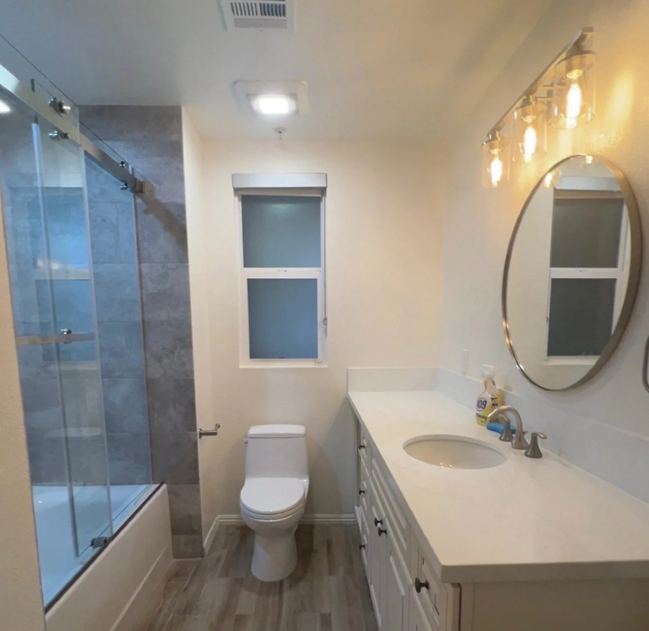 Bathroom with glass-enclosed shower, toilet, white vanity with sink, oval mirror, frosted window, and wood-look flooring.