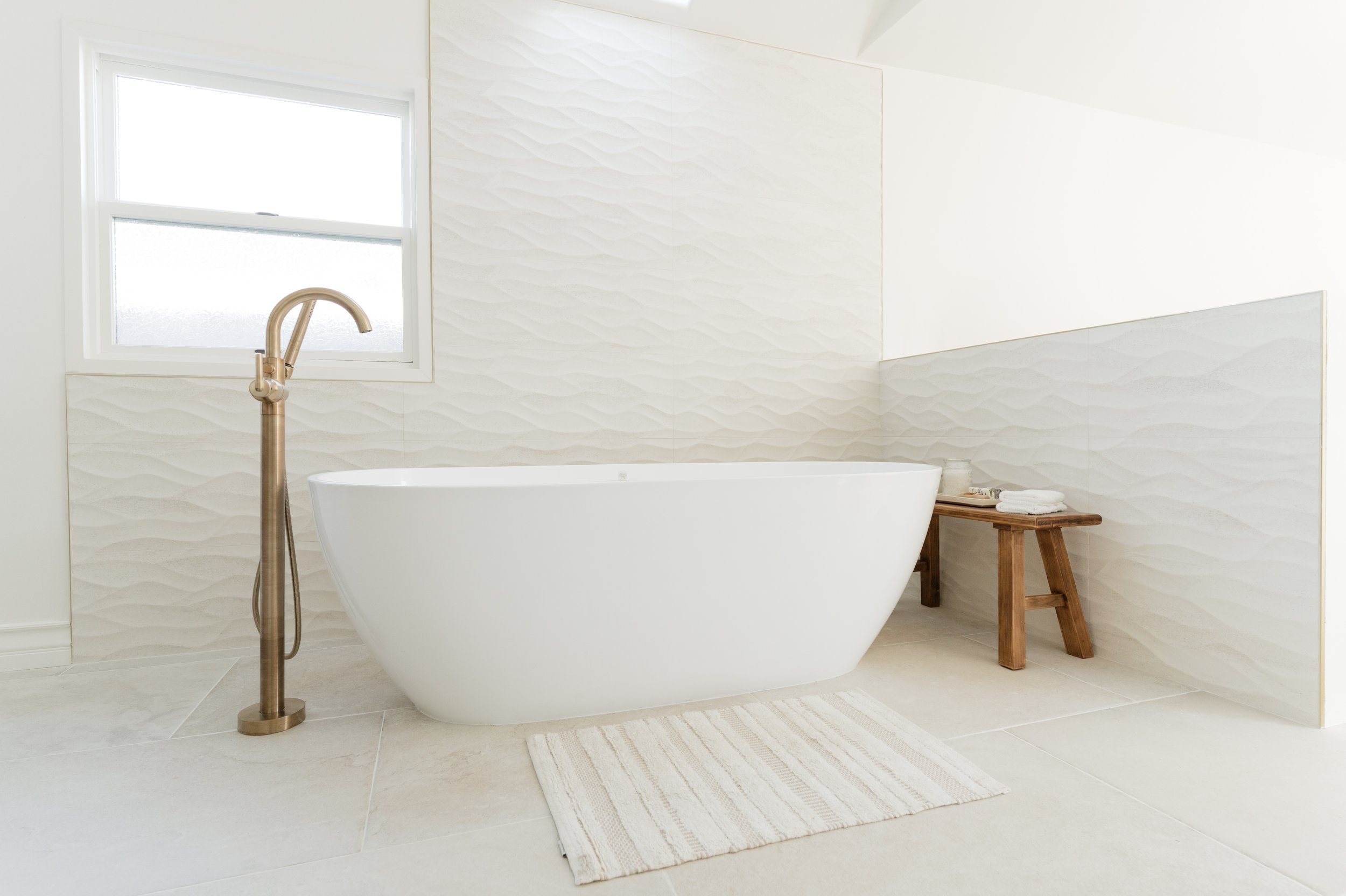 A modern bathroom with a freestanding bathtub, wooden bench with towels, a small window, and textured white tiles.
