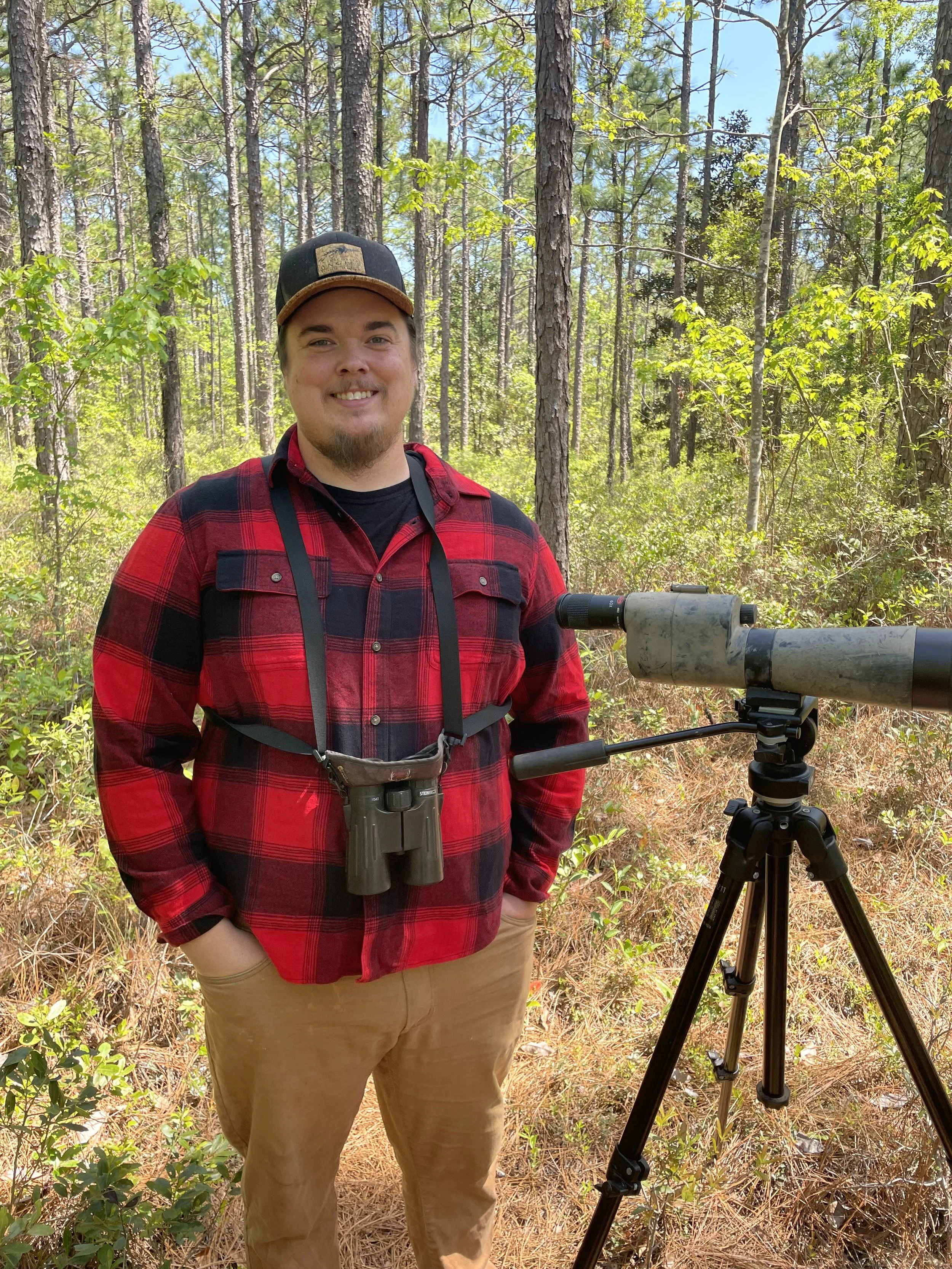 Person in plaid shirt with hat and binoculars standing in a forest next to a telescope on a tripod.