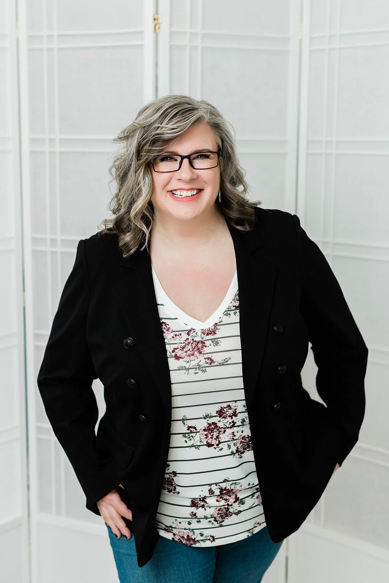 A smiling woman with gray, wavy hair and glasses, wearing a black blazer over a floral and striped top, standing in front of a white folding screen.