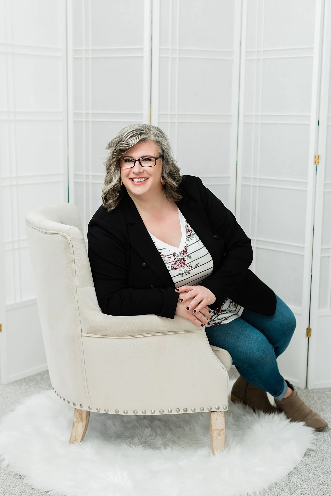 Person sitting on a beige chair, wearing glasses, a black blazer, jeans, and a patterned top, smiling.