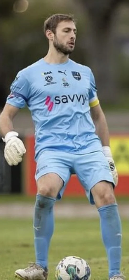Soccer goalkeeper in light blue uniform with gloves, standing near a soccer ball on a field.