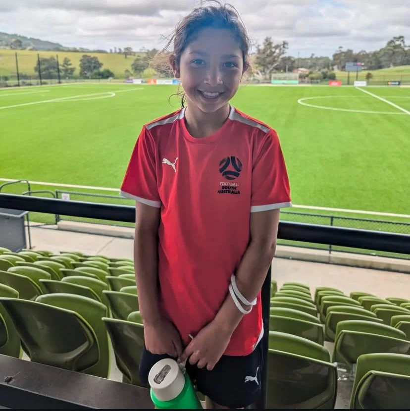 Girl in red soccer jersey standing in front of an empty sports field with green grass and bleachers.