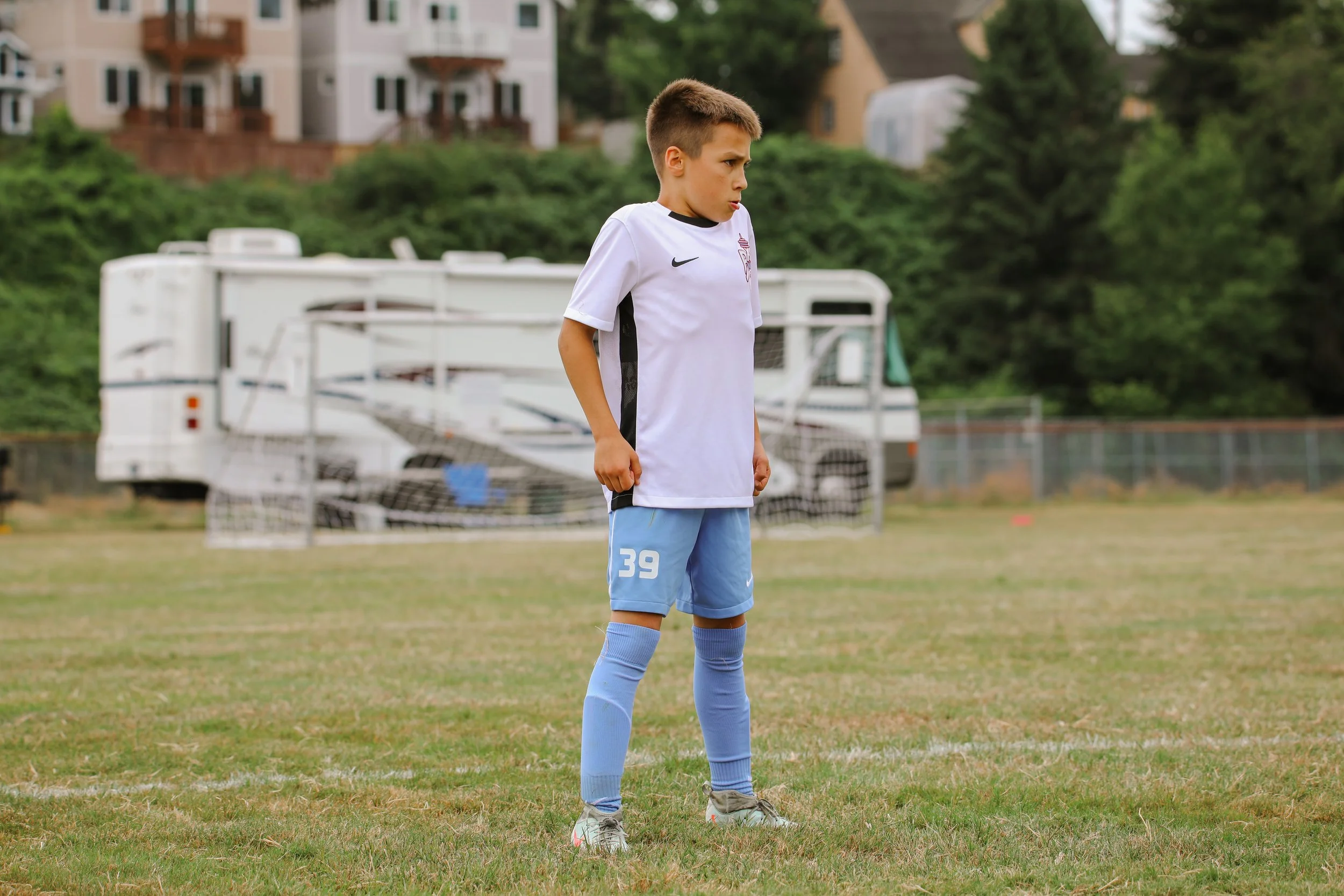 A young boy standing on a soccer field wearing a white soccer jersey, blue shorts with the number 39, and blue socks, with a soccer goal and a recreational vehicle in the background.