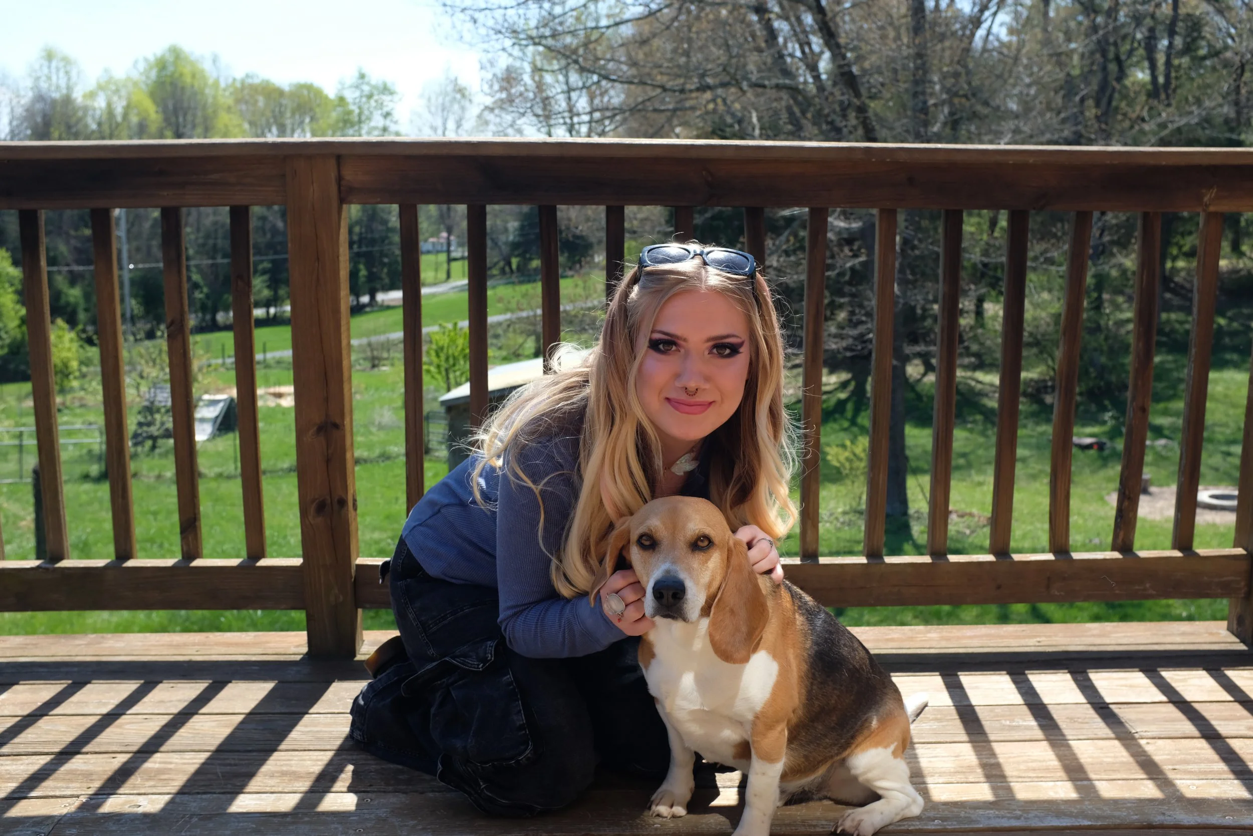 A woman with blonde hair, sunglasses on her head, and makeup crouching next to a beagle dog outdoors on a wooden deck with a green backyard and trees in the background.