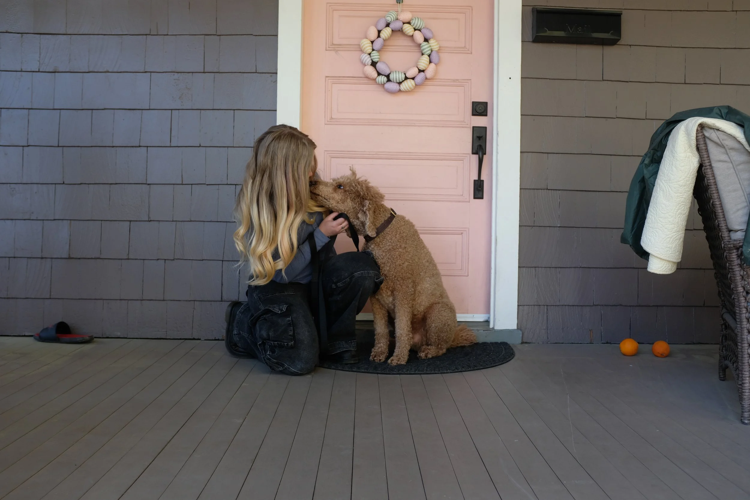 A young girl kneeling on a porch, kissing a brown curly-haired dog sitting on a black doormat in front of a pink door with a decorative wreath. There are two oranges on the ground to the right and a pair of sandals to the left.
