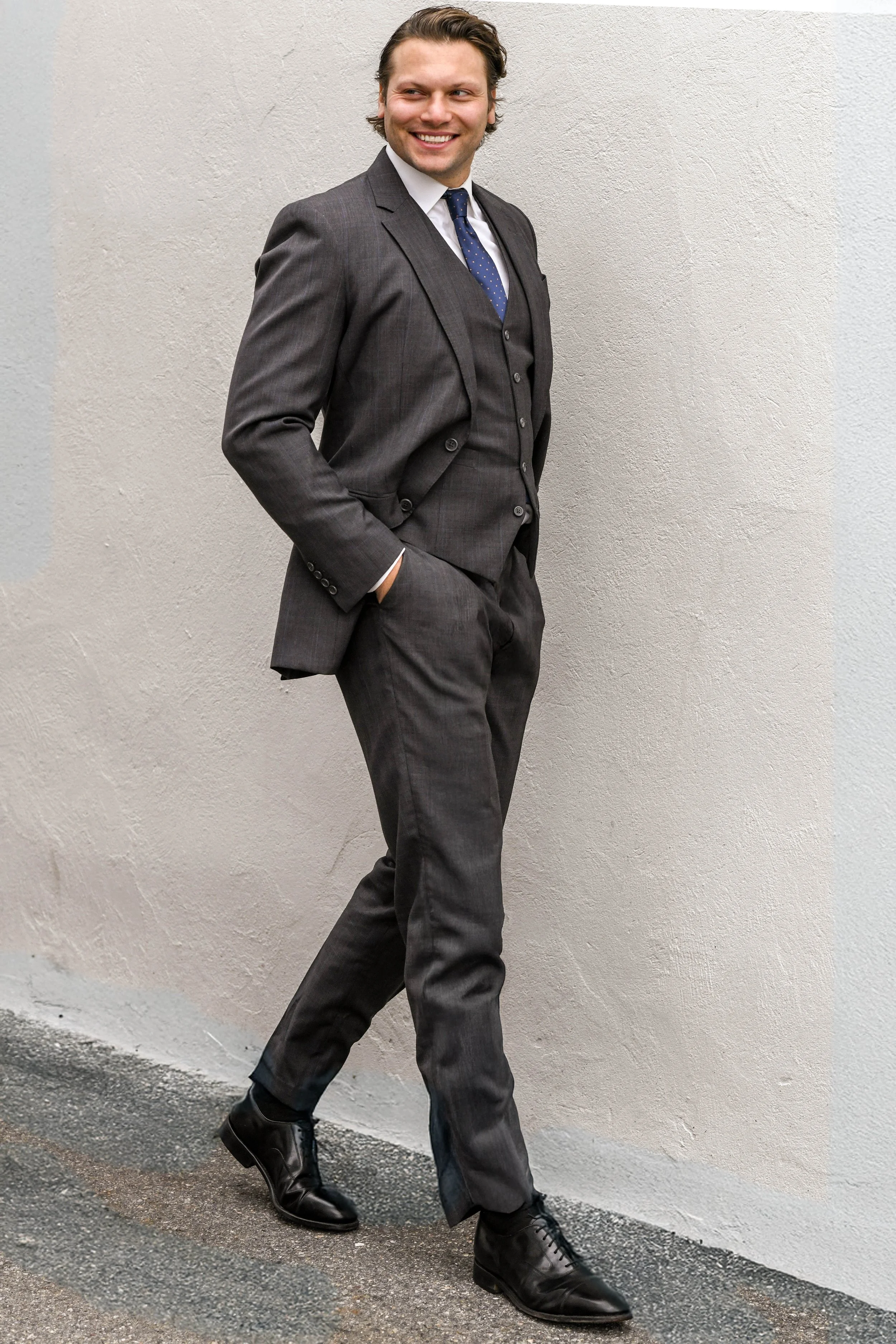 Ben Forman in a dark gray suit, white shirt, and blue polka-dot tie, standing against a light-colored wall with hands in his pockets.