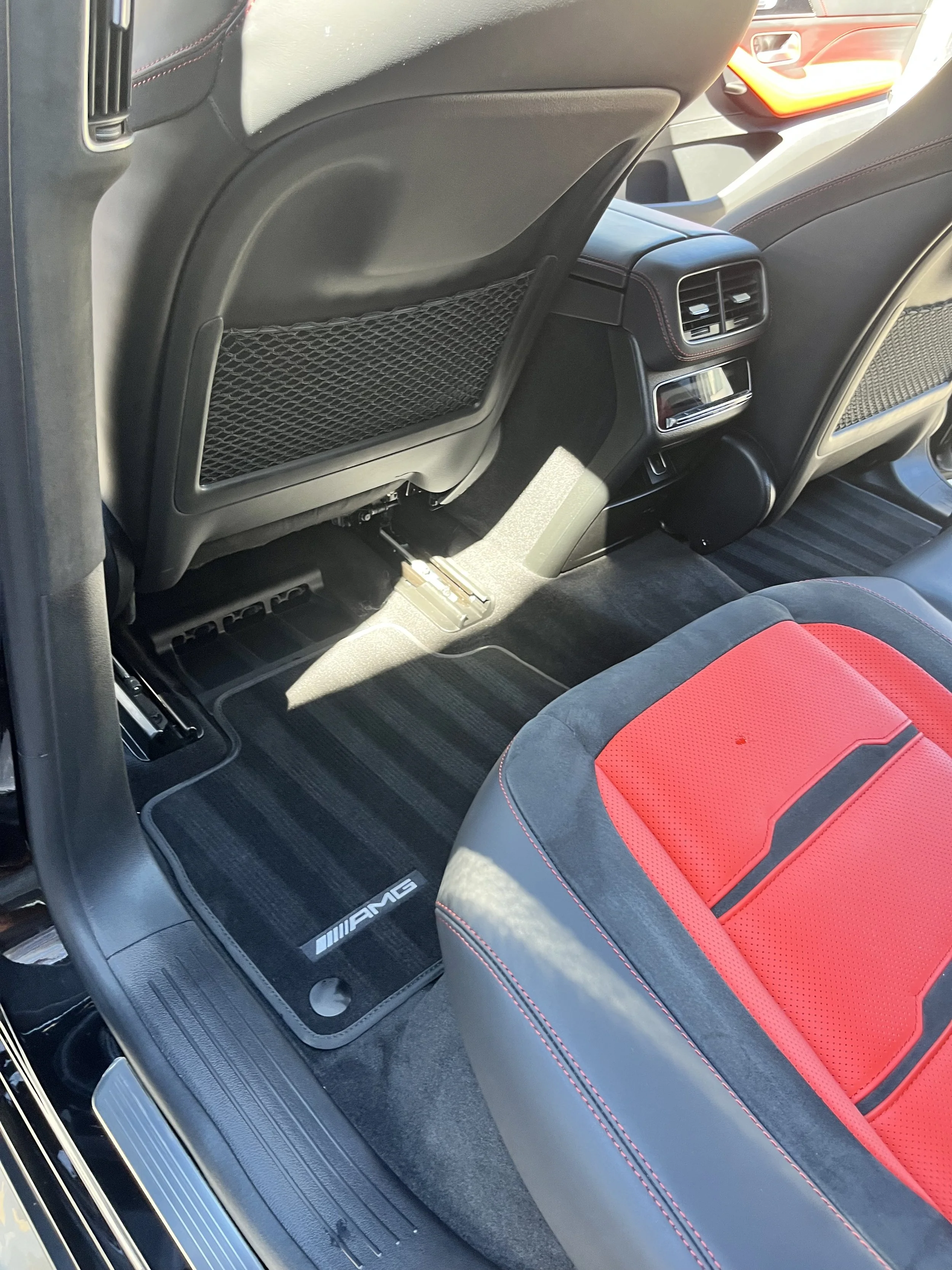 The interior of a car showing the backseat area with black and red seat covers, AMG branded floor mats, and back of front seats with storage pockets.