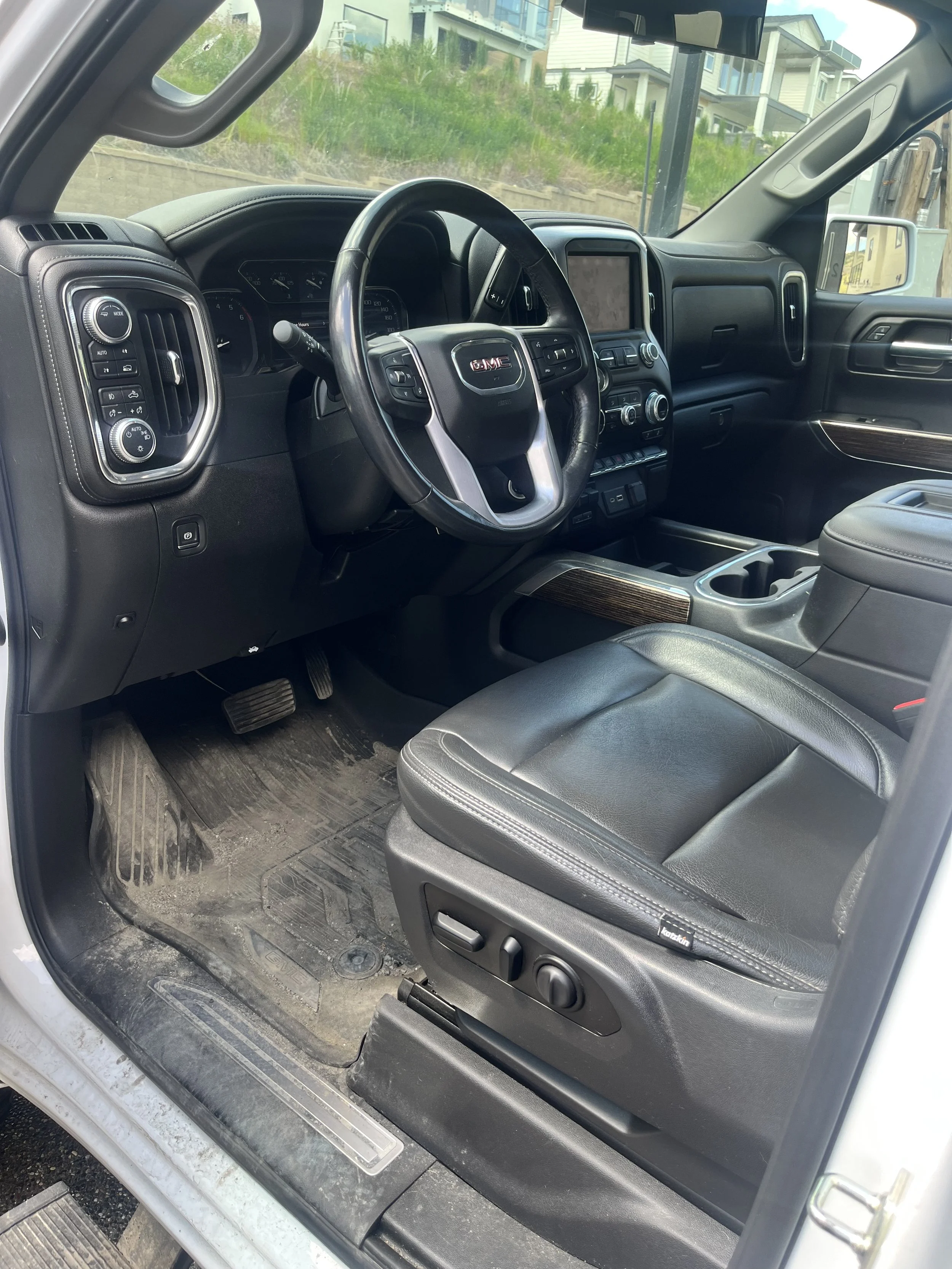 Interior of a GMC truck showcasing the driver’s seat, dashboard, and steering wheel, with visible control buttons, cup holders, and a GPS screen, parked outdoors near houses.