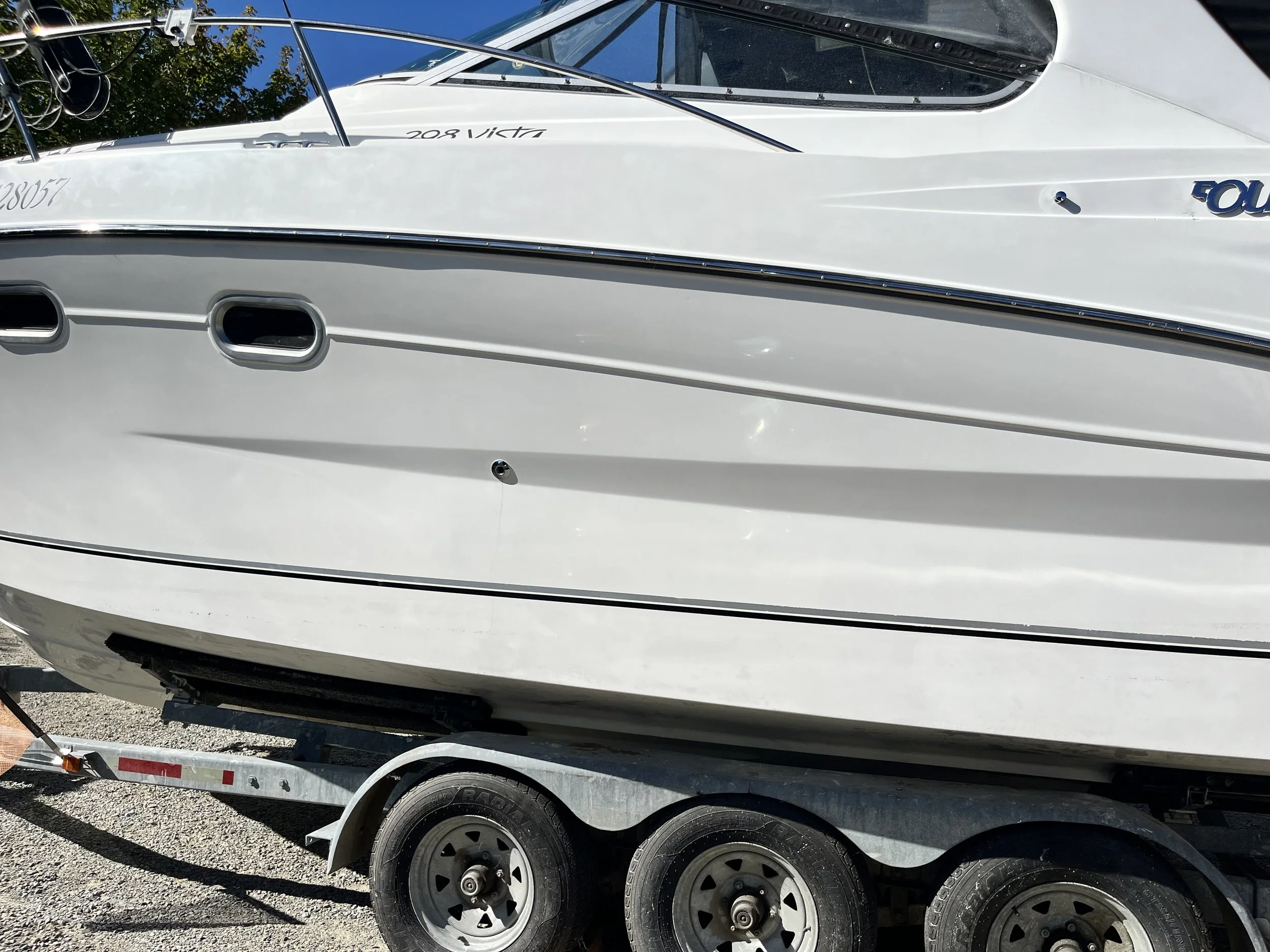 A white boat on a trailer with black tires, parked on gravel, with some trees and blue sky reflected on the boat's surface.