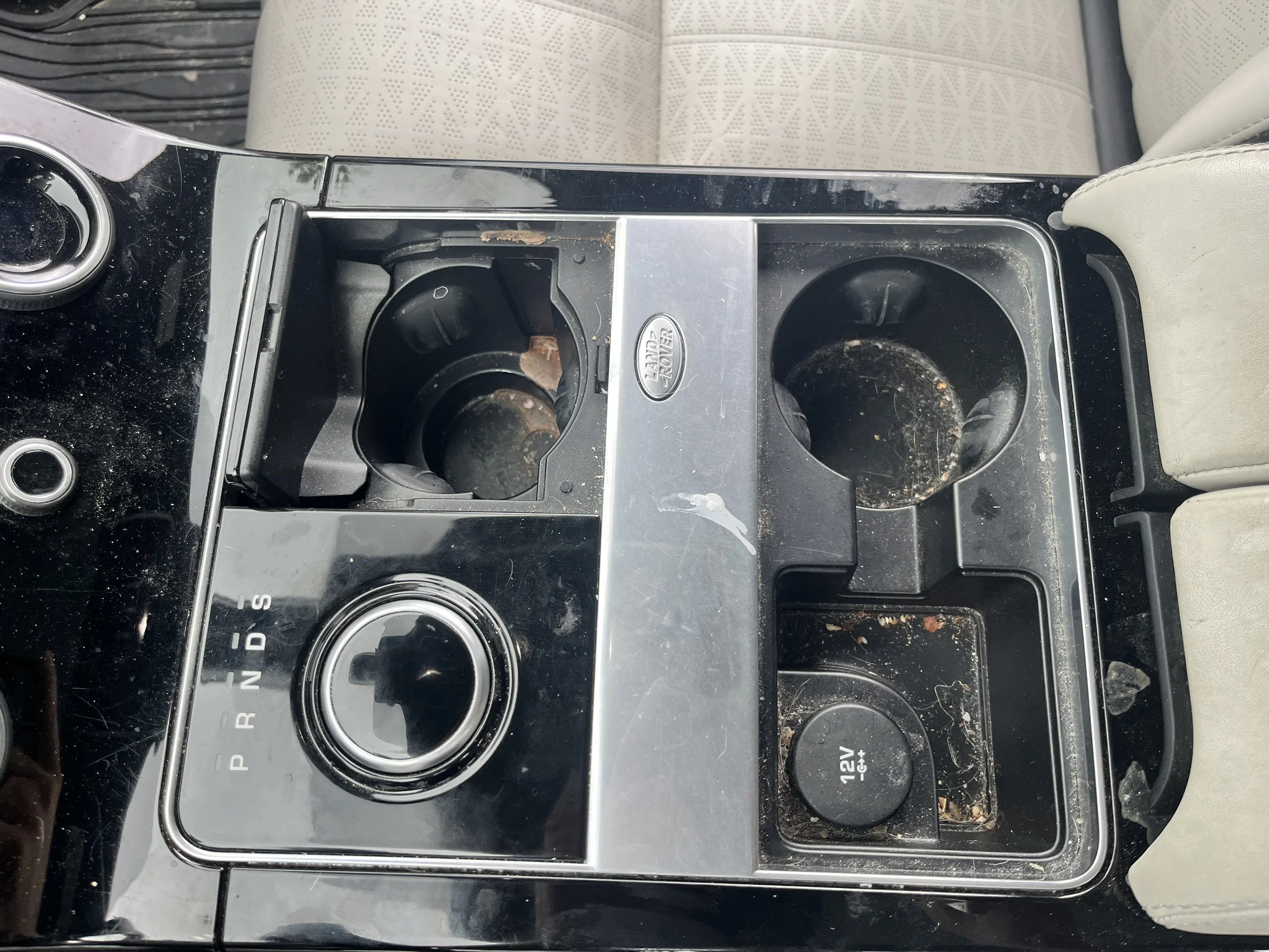 Dirty car center console with cup holders, gear shift, 12V power outlet, and various debris.