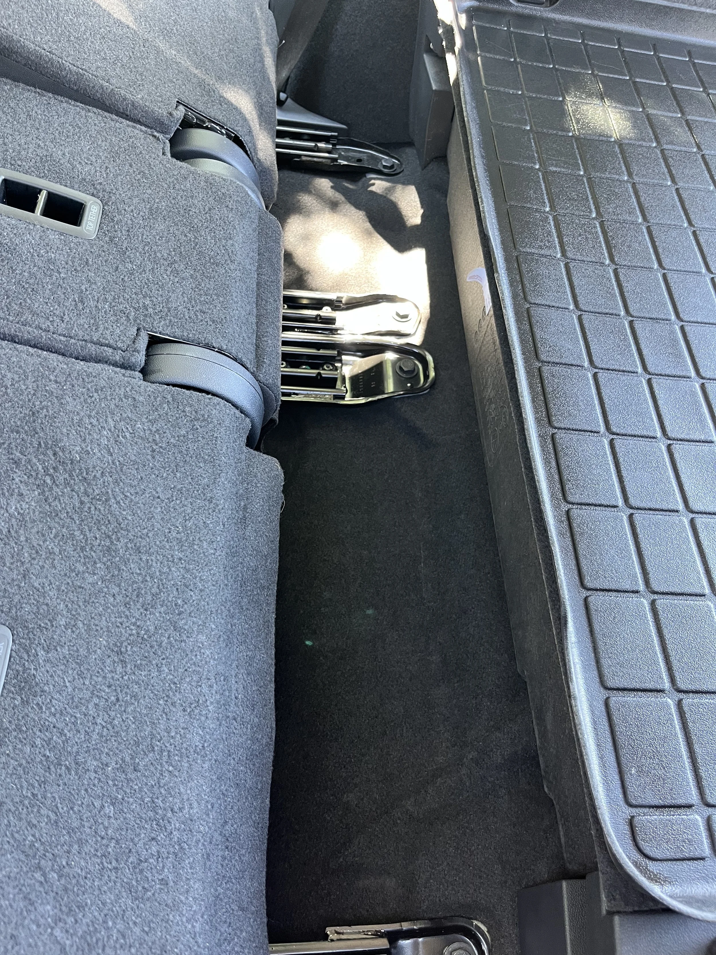 The backseat of a vehicle showing folded metal scooter or wheelchair frames, a black rubber floor mat, and part of the vehicle's carpeted interior.