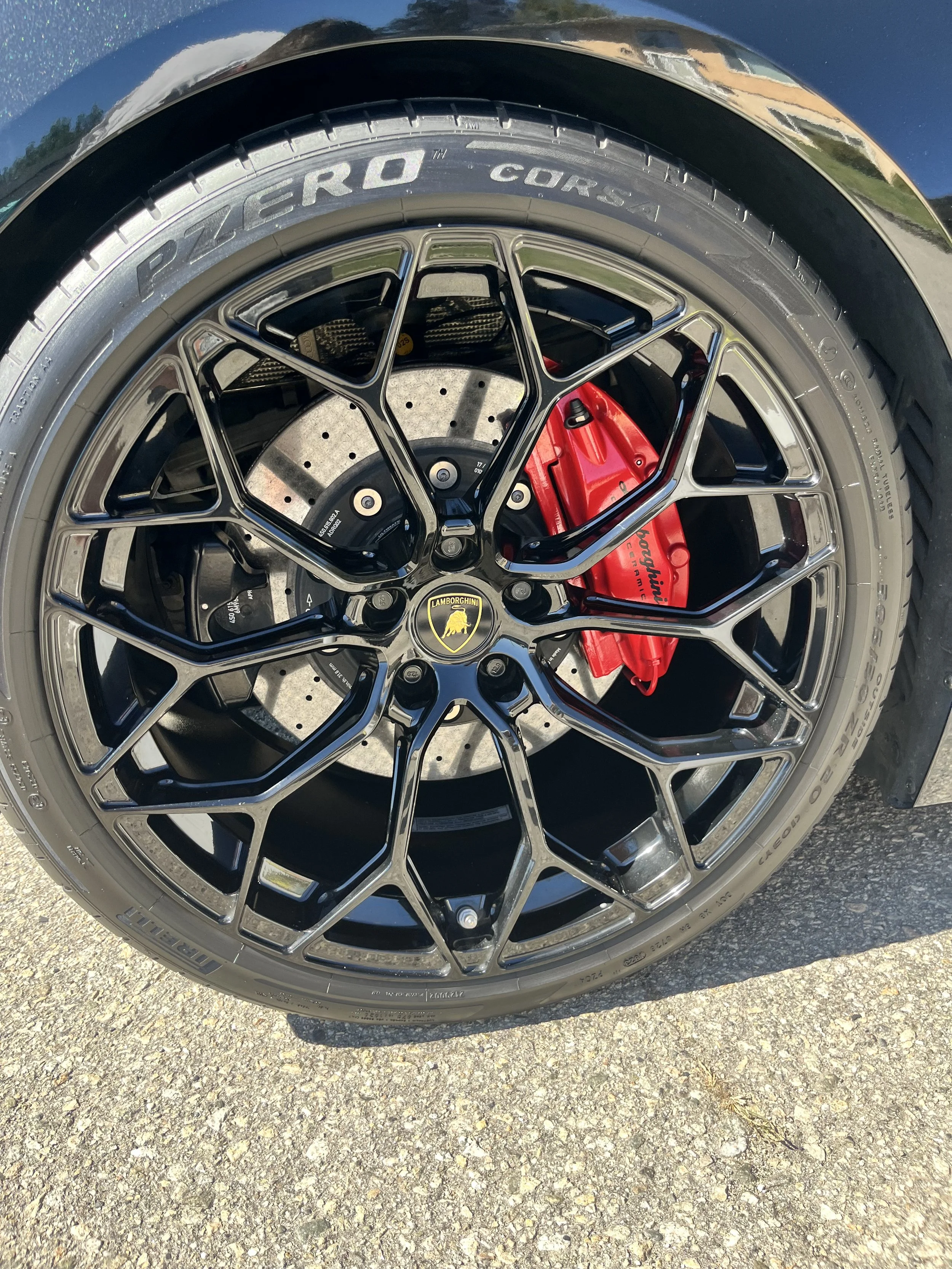 Close-up of a car tire with the wheel rim and brake caliper visible, featuring a Lamborghini logo on the center cap and a red brake caliper behind the rim.