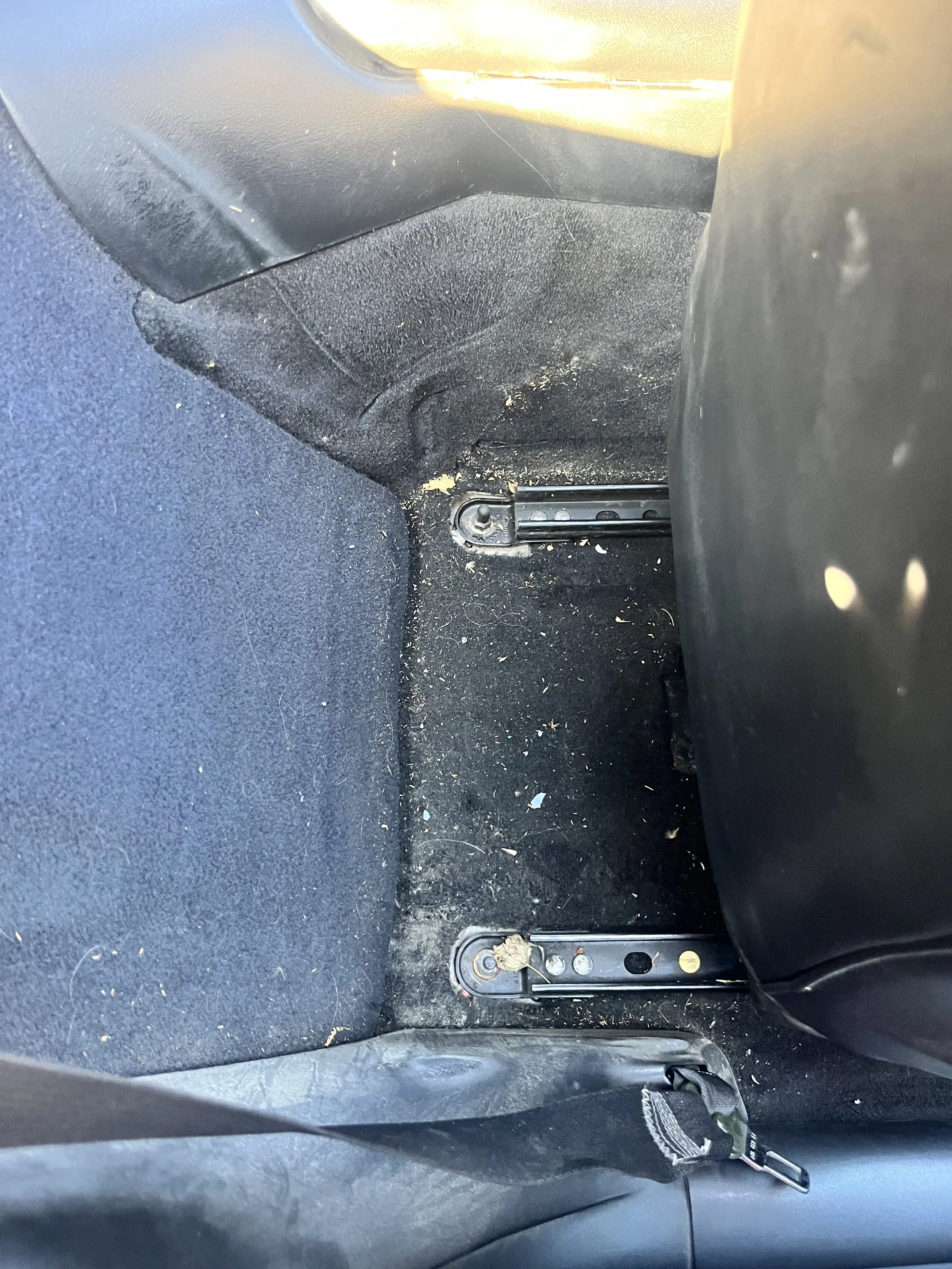 Car backseat with the front seat removed, showing the metal rails and floor carpet, which is dirty with small debris and dust.