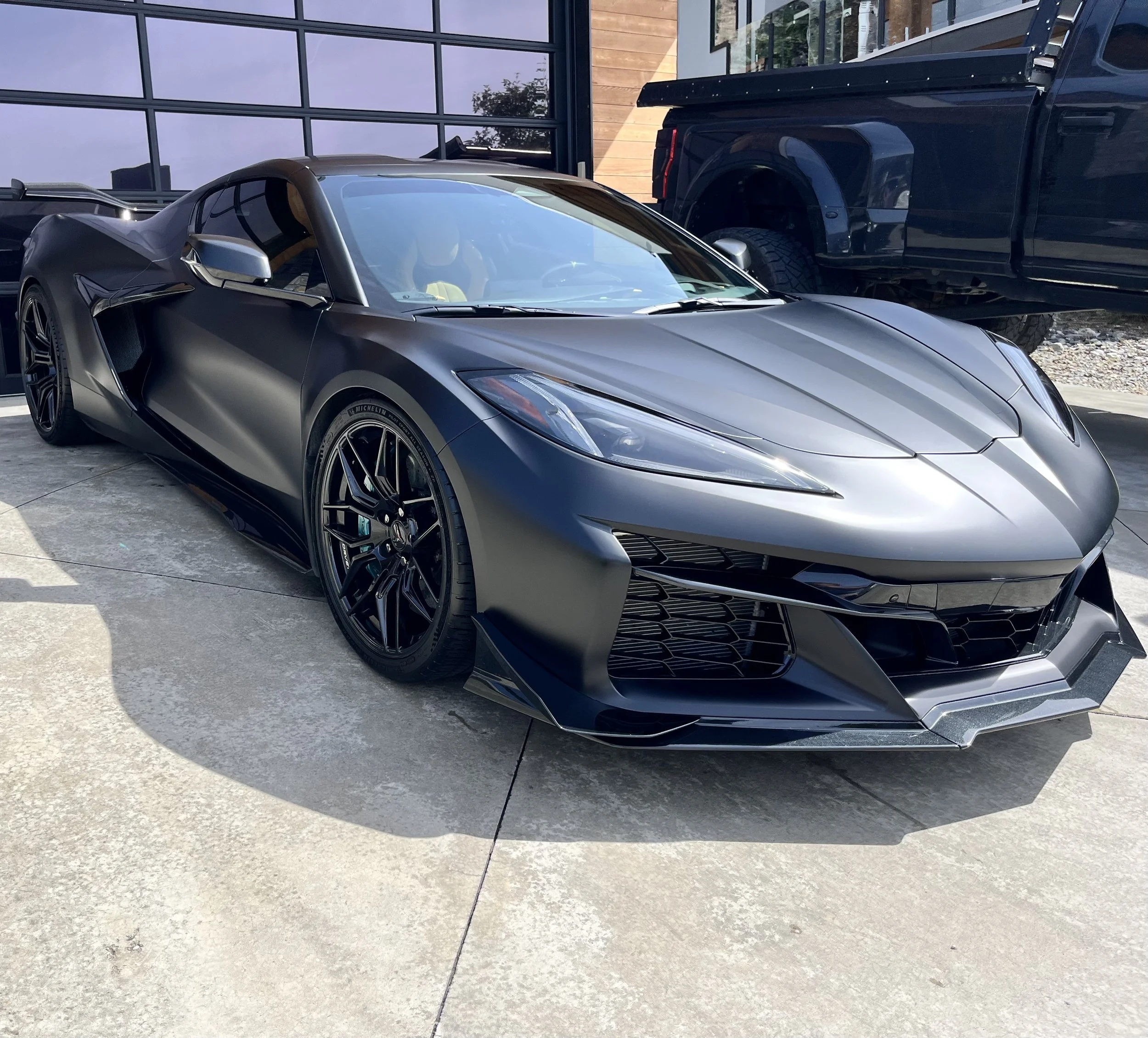 A dark gray sports car with sleek design, black wheels, and aerodynamic features parked on a concrete driveway outside a modern building with large windows.