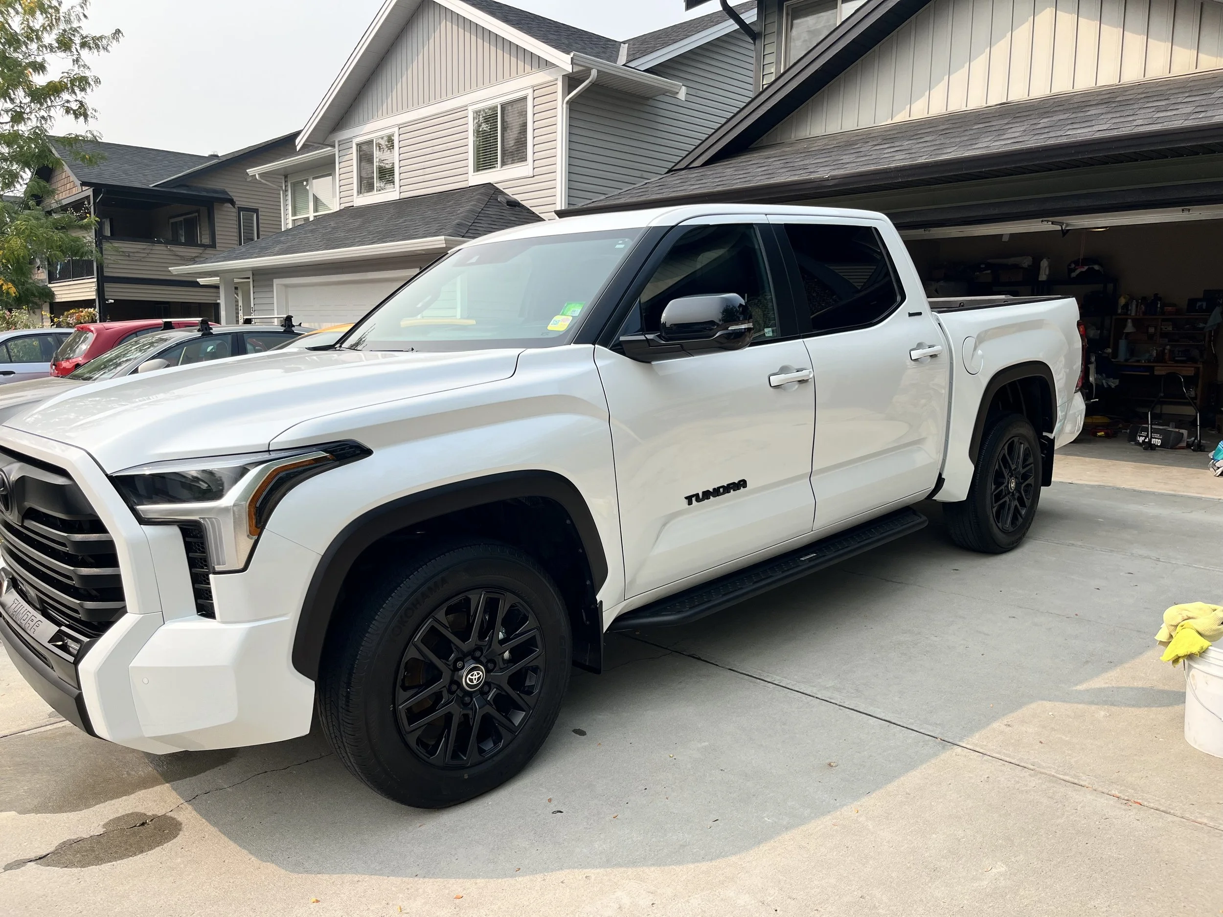 White Toyota Tundra pickup truck parked in a driveway in front of a garage, with houses and other vehicles in the background.