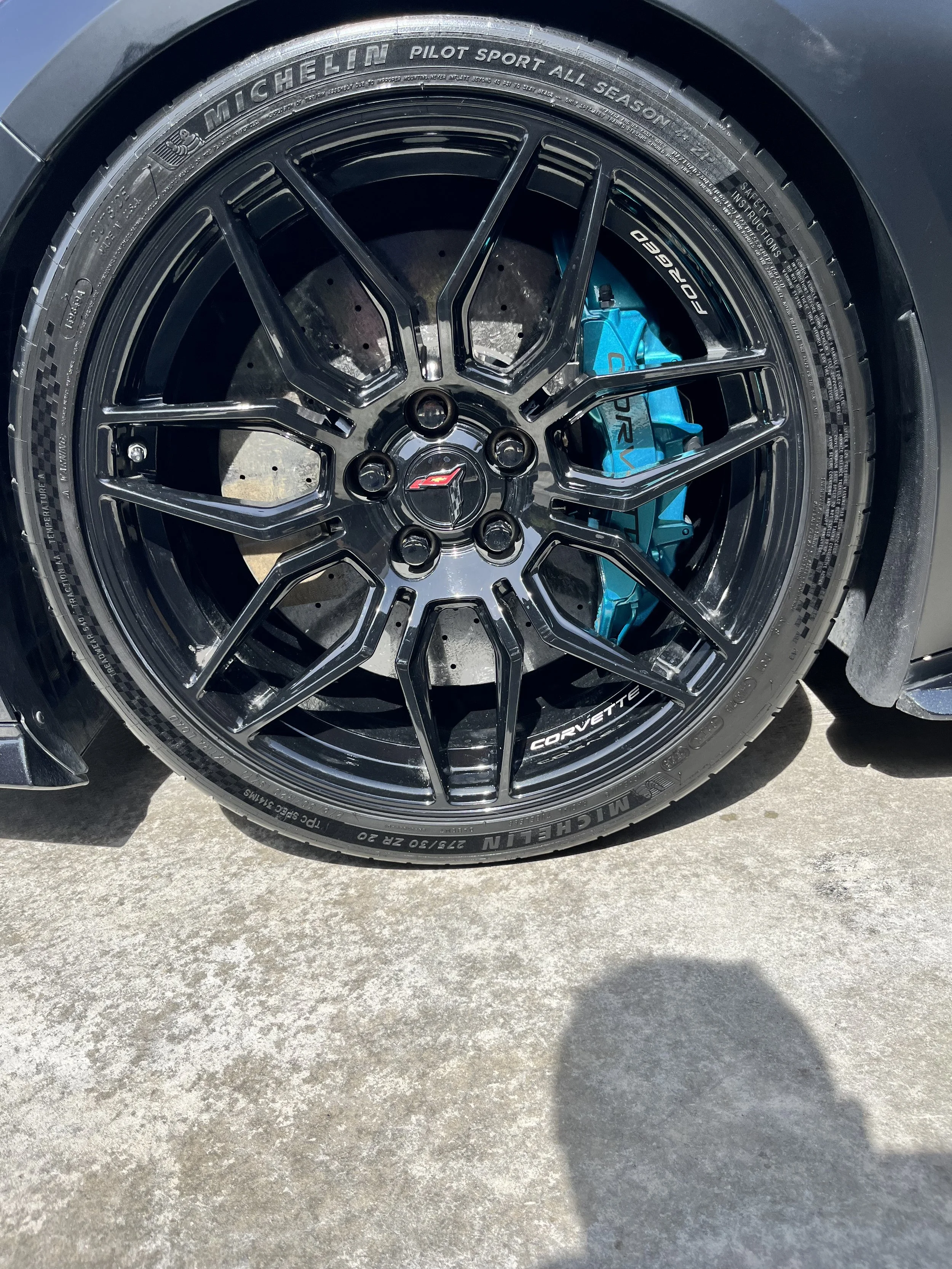 Close-up of a black alloy wheel with blue brake caliper behind it, mounted with Michelin Pilot Sport tires, on a Corvette.
