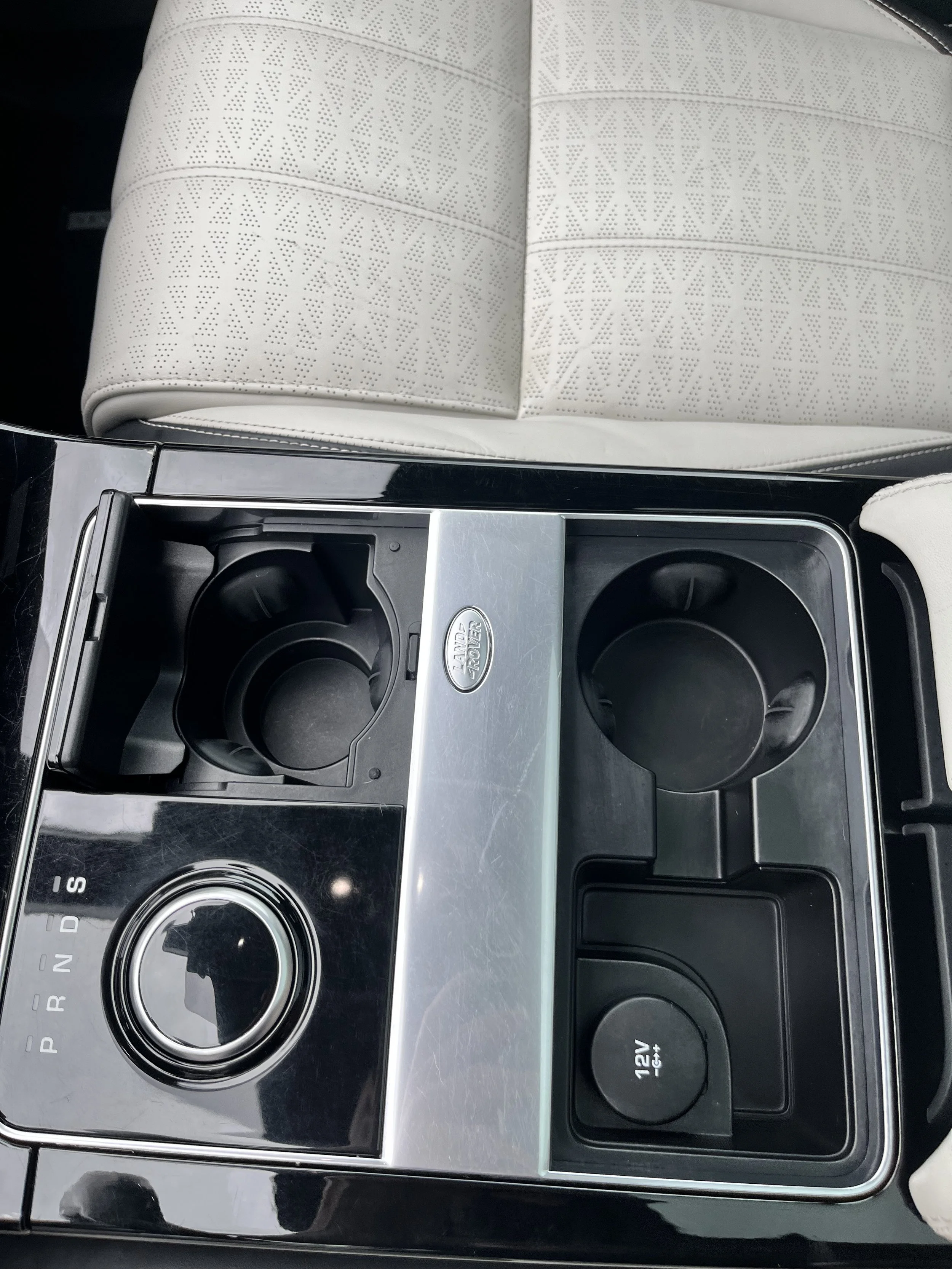 Car center console with two cup holders, a rotary gear shift, and a 12V power outlet. Part of a white leather car seat is visible above the console.
