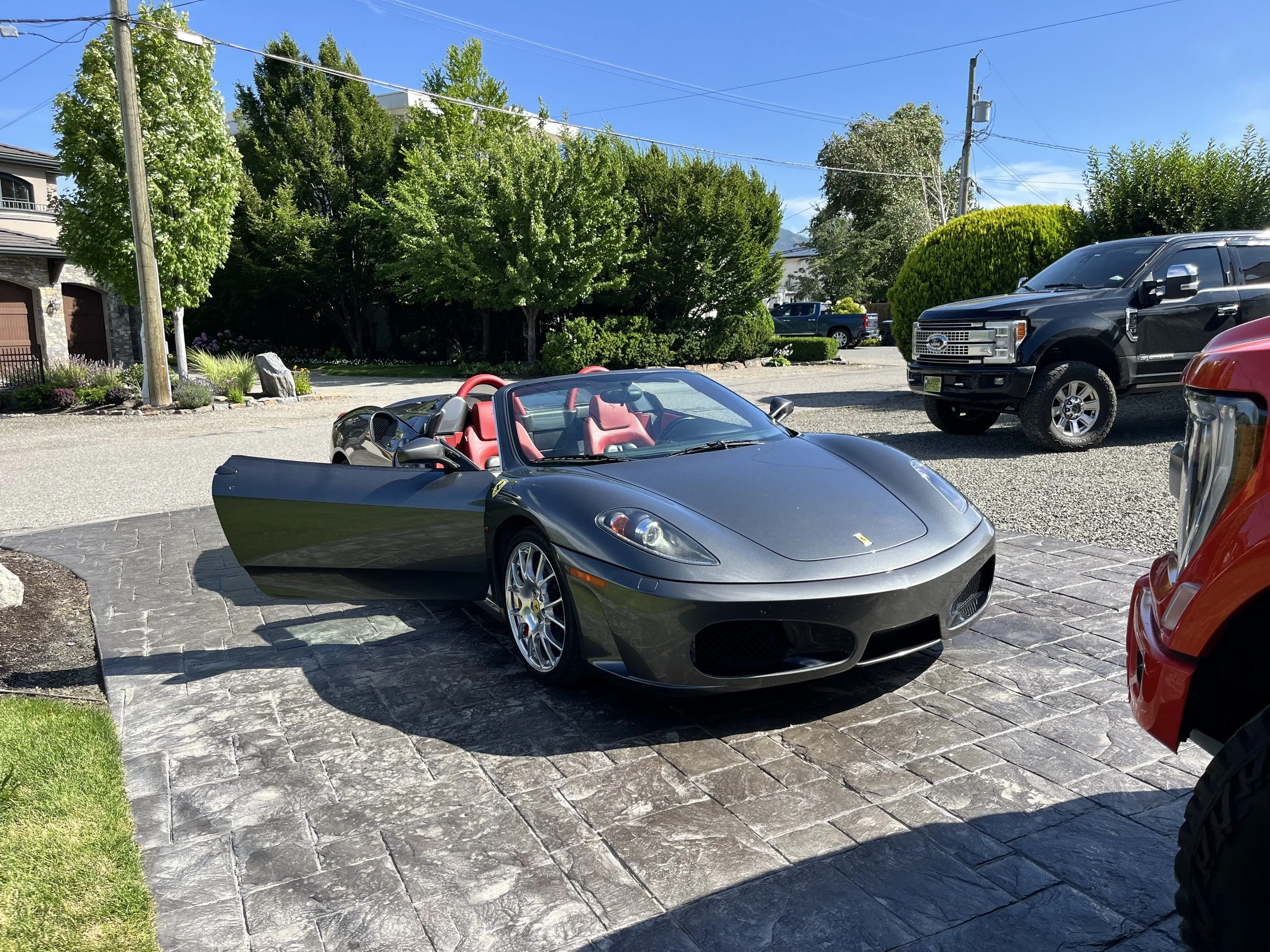 A silver Ferrari convertible with pink interior parked on a stone driveway, with a black truck and a red vehicle nearby under a clear blue sky.