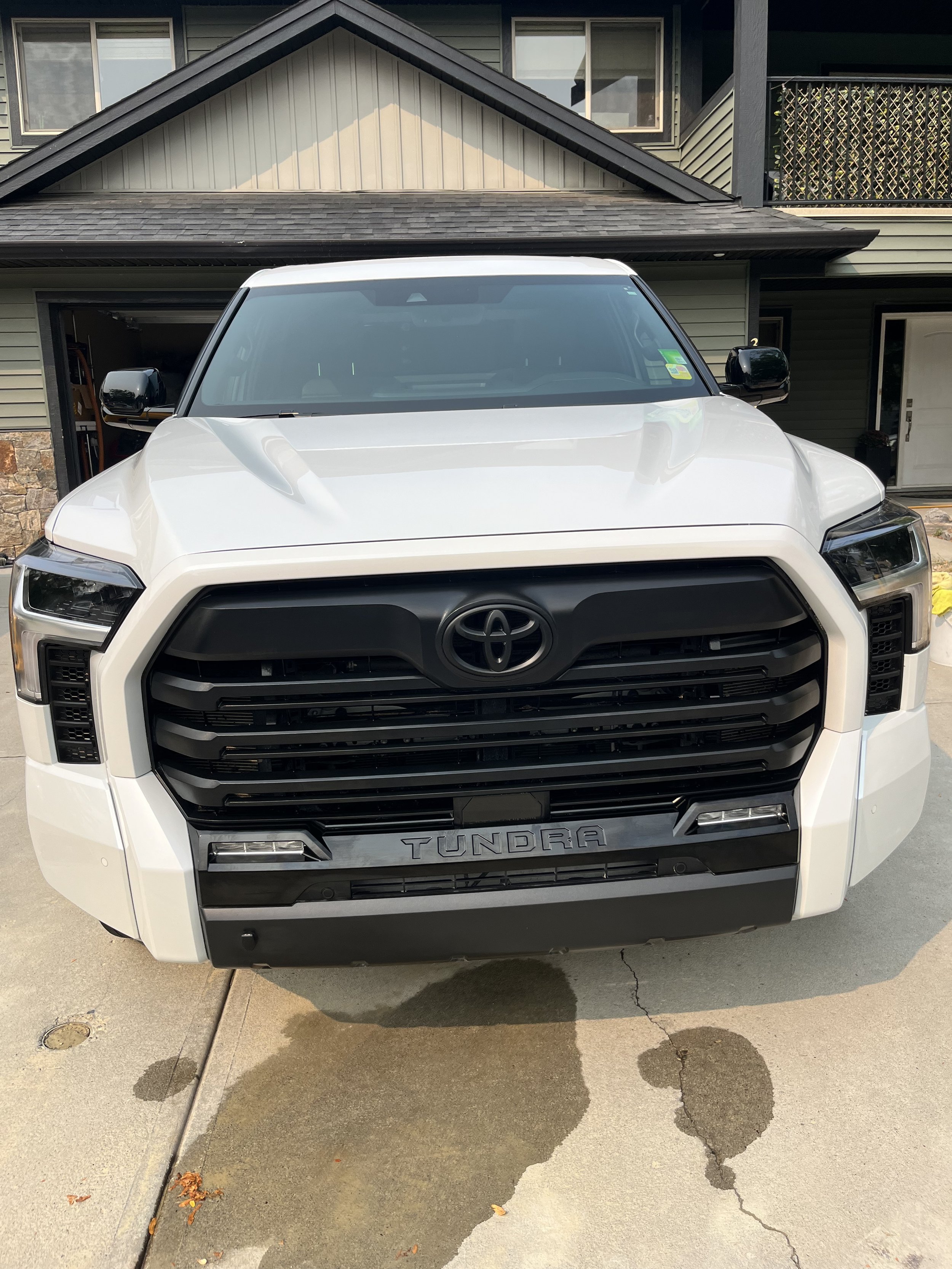 Front view of a white Toyota Tundra pickup truck parked on concrete driveway in front of a modern house with a garage, stone accents, and multiple stories.