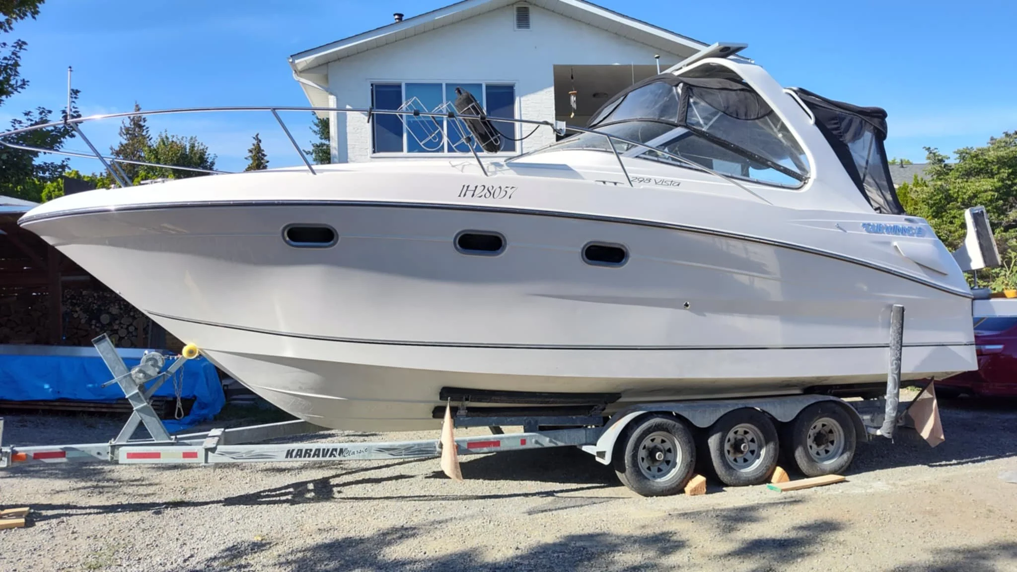 A white boat on a trailer parked outside in a residential area near a house, with trees and a clear blue sky in the background.