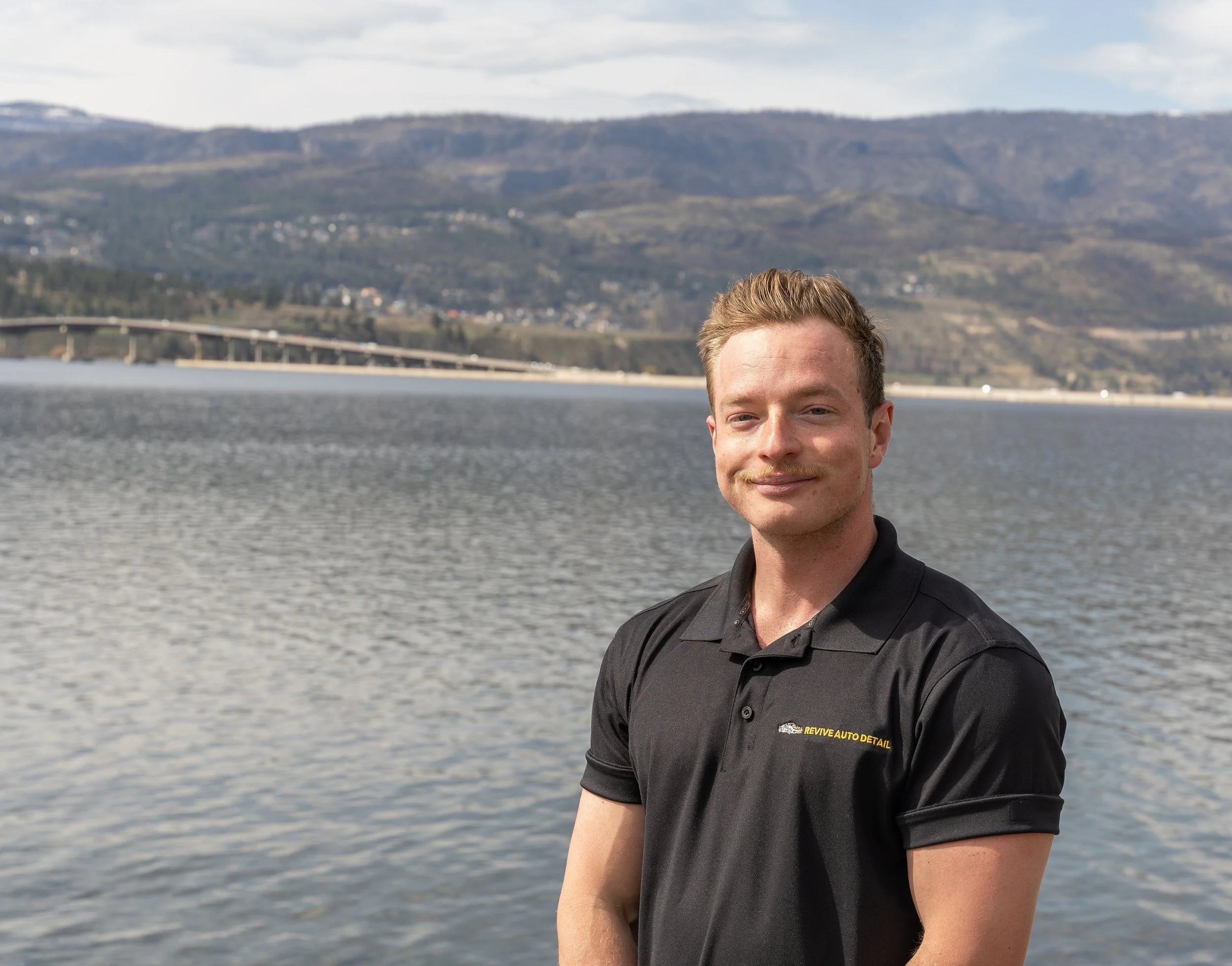 A man with light skin, short light brown hair, and a slight mustache and beard, standing outdoors by a body of water with mountains in the background, wearing a black polo shirt with yellow and white text.