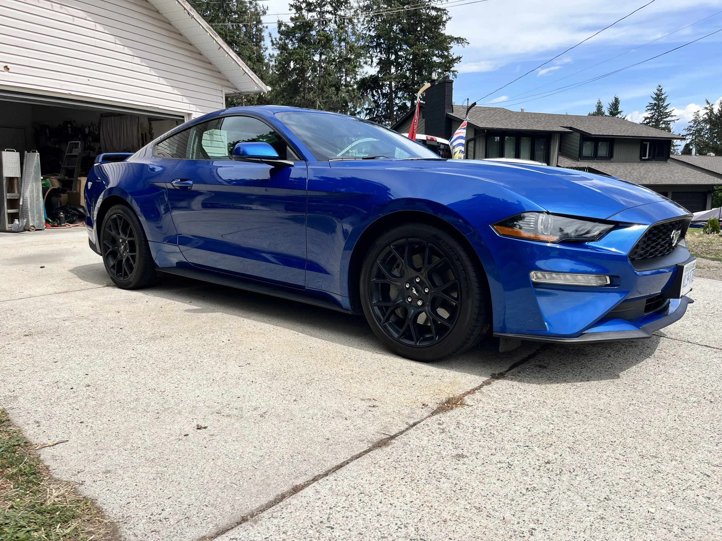 A blue sports car parked on a driveway in front of a house with an open garage, suburban neighborhood with trees and houses in the background.