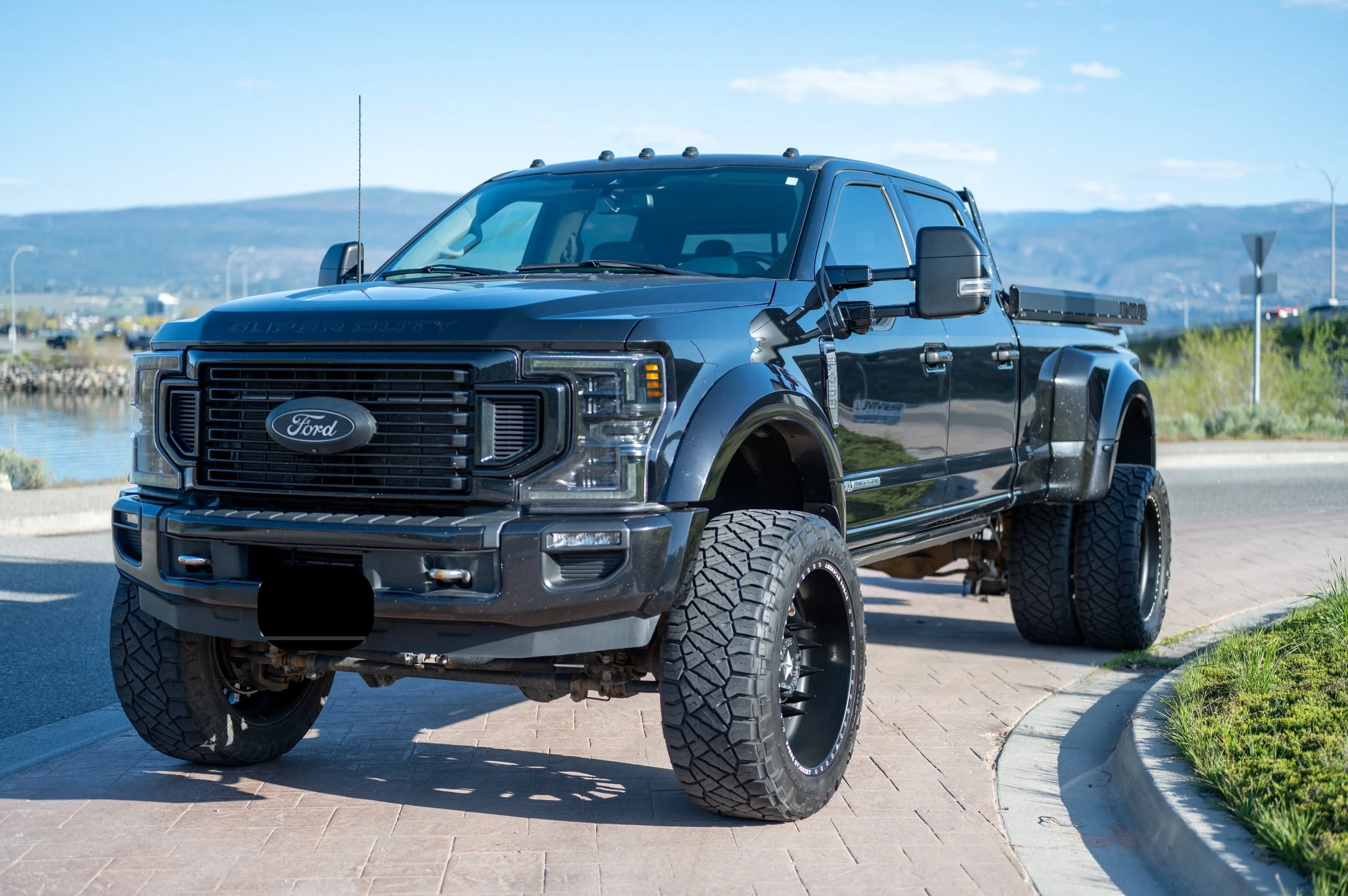 Black lifted Ford pickup truck parked on a street near water with hilly landscape in the background.
