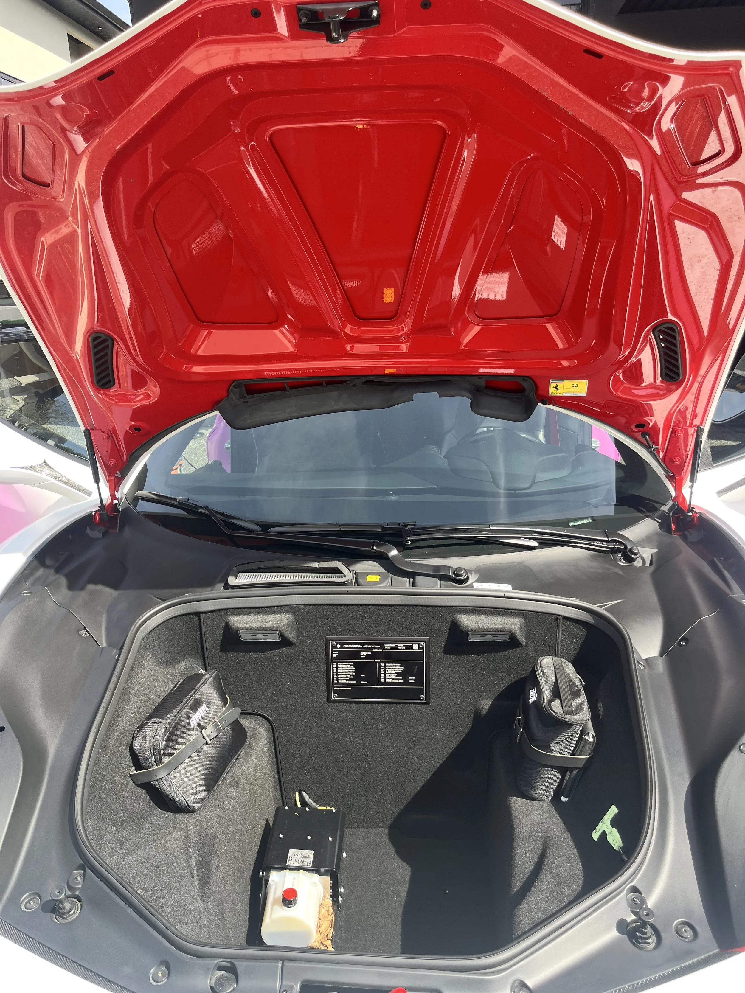 Front trunk of a red sports car with open hood, showing storage area with a small black bag on the left, electronic component in the middle, and a small black bag on the right.