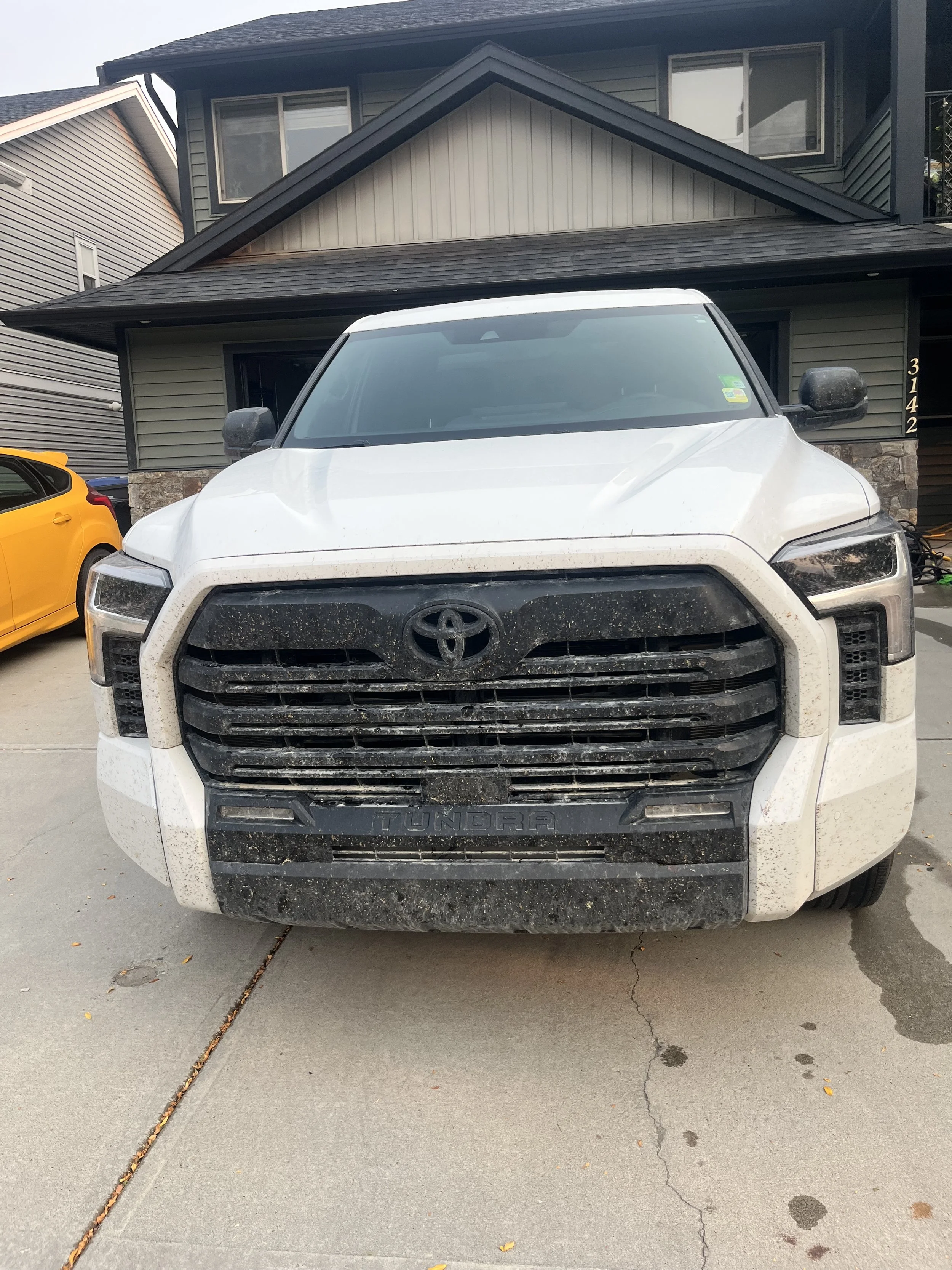 Front view of a dusty white Toyota Tundra truck parked on a concrete driveway in front of a house with dark green siding and a black roof, with a yellow car partially visible on the left side.