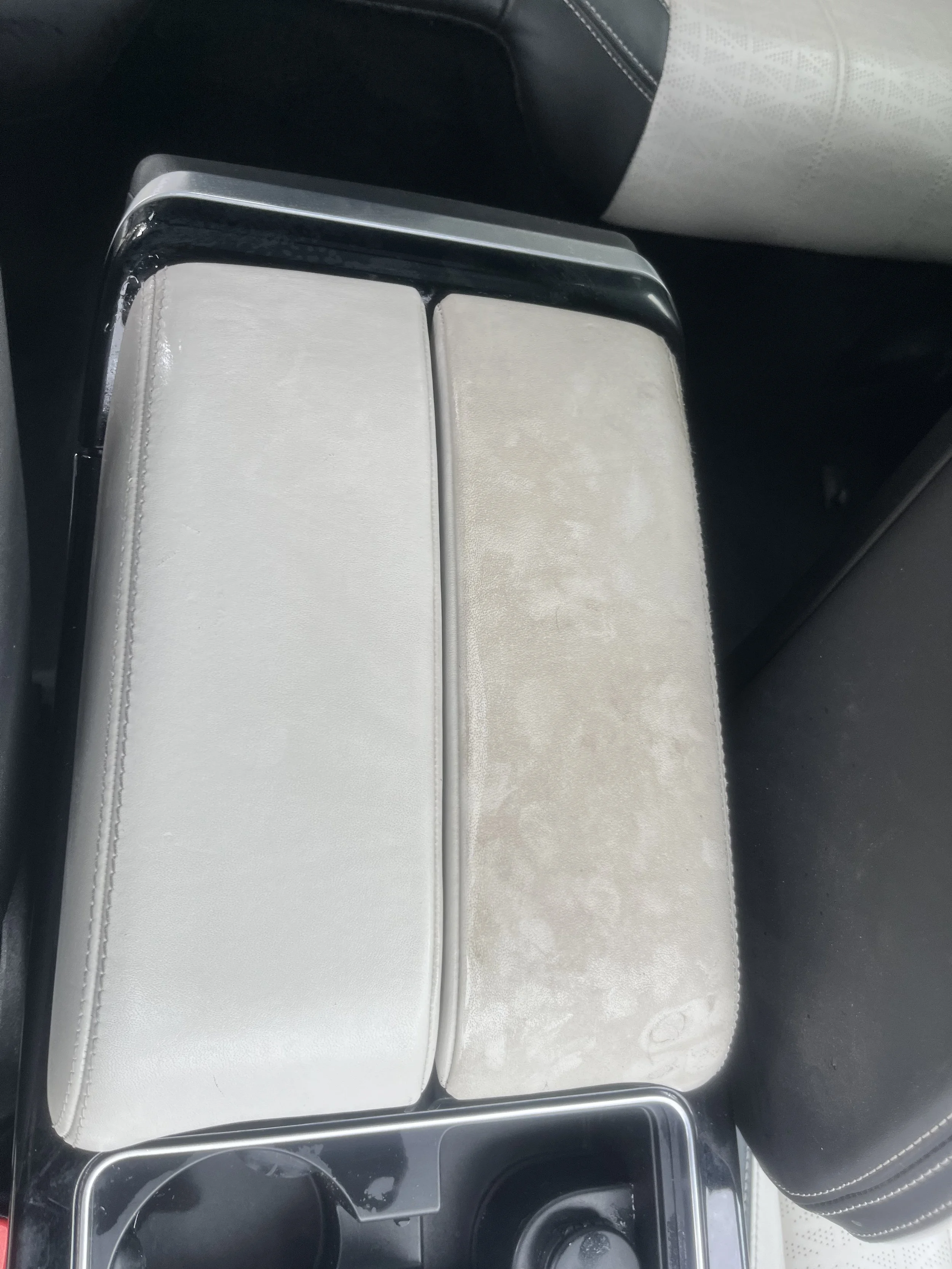 Interior view of a vehicle's center console with two closed compartments, one covered in white leather and the other in light-colored suede or fabric, with a cup holder in the lower left corner.