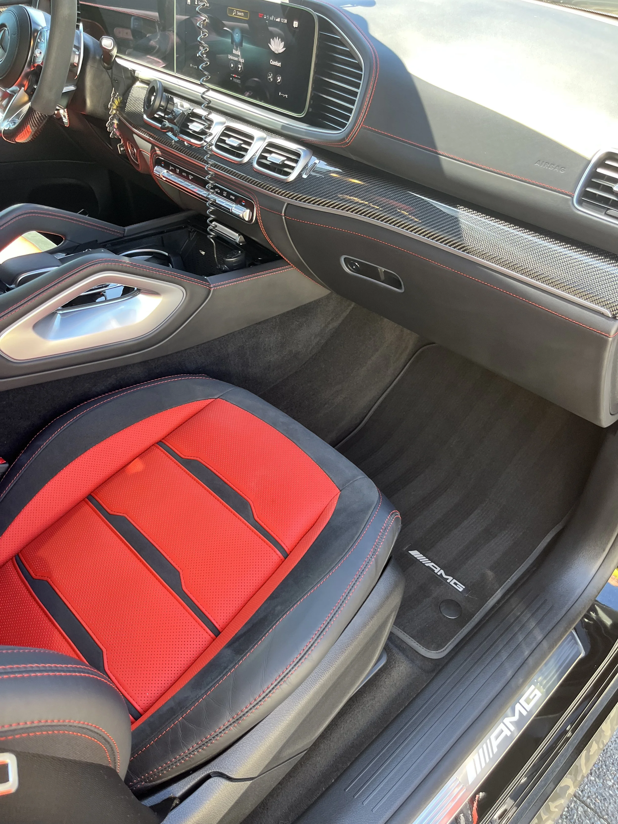 The interior of a high-performance Mercedes-AMG vehicle featuring a red and black racing-style seat, a digital dashboard display, carbon fiber accents, and AMG branding on the floor mat.