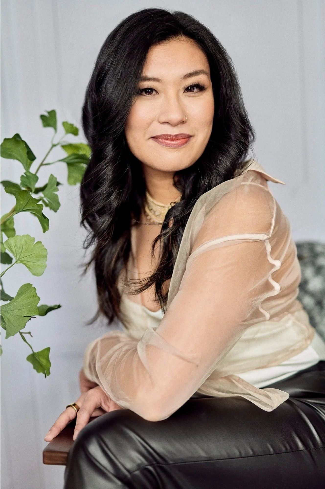 Woman with long black hair wearing a sheer blouse and leather pants, sitting near a plant with a neutral expression.