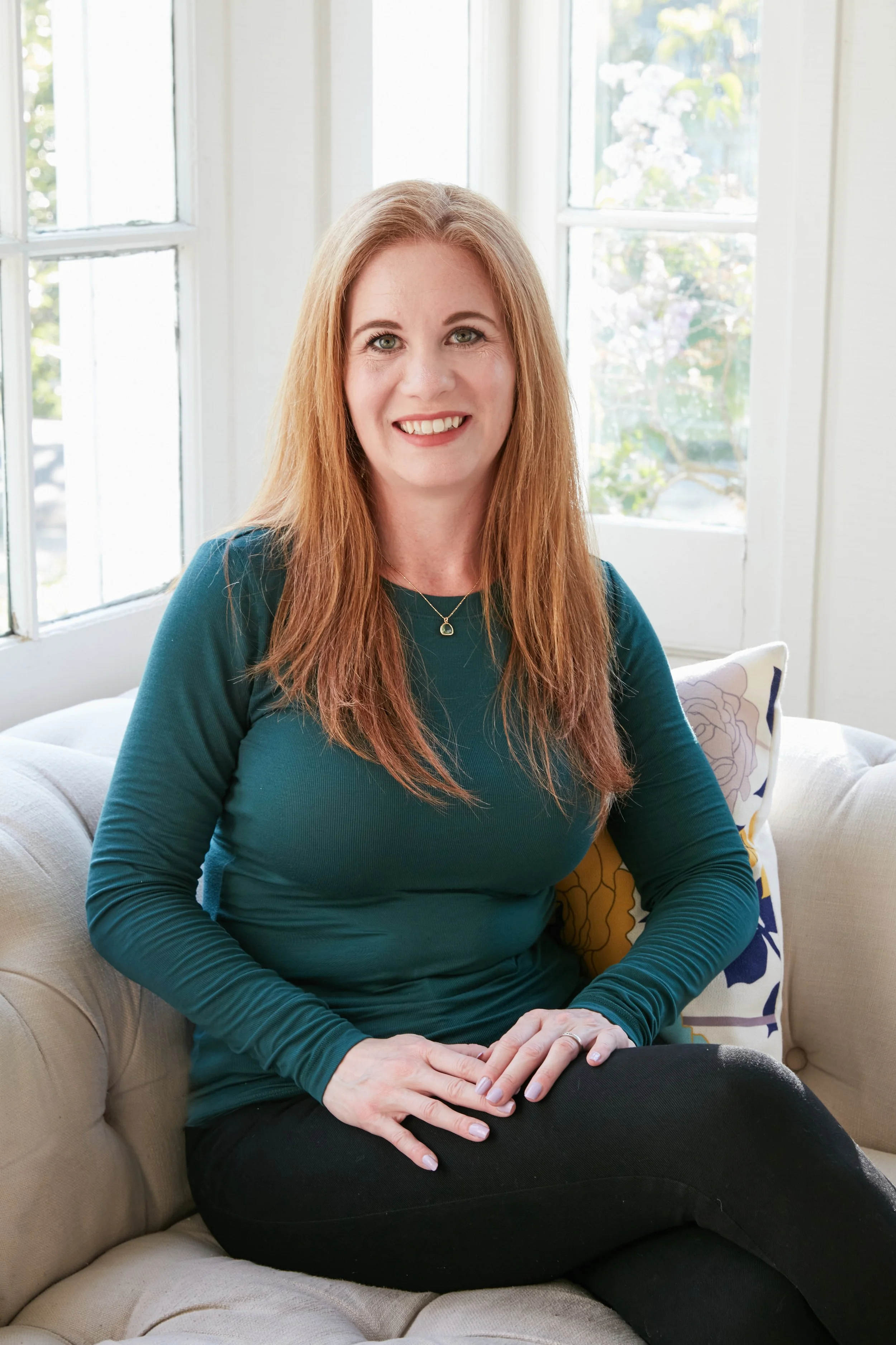Woman with long red hair in green top sitting on a sofa in a bright room.