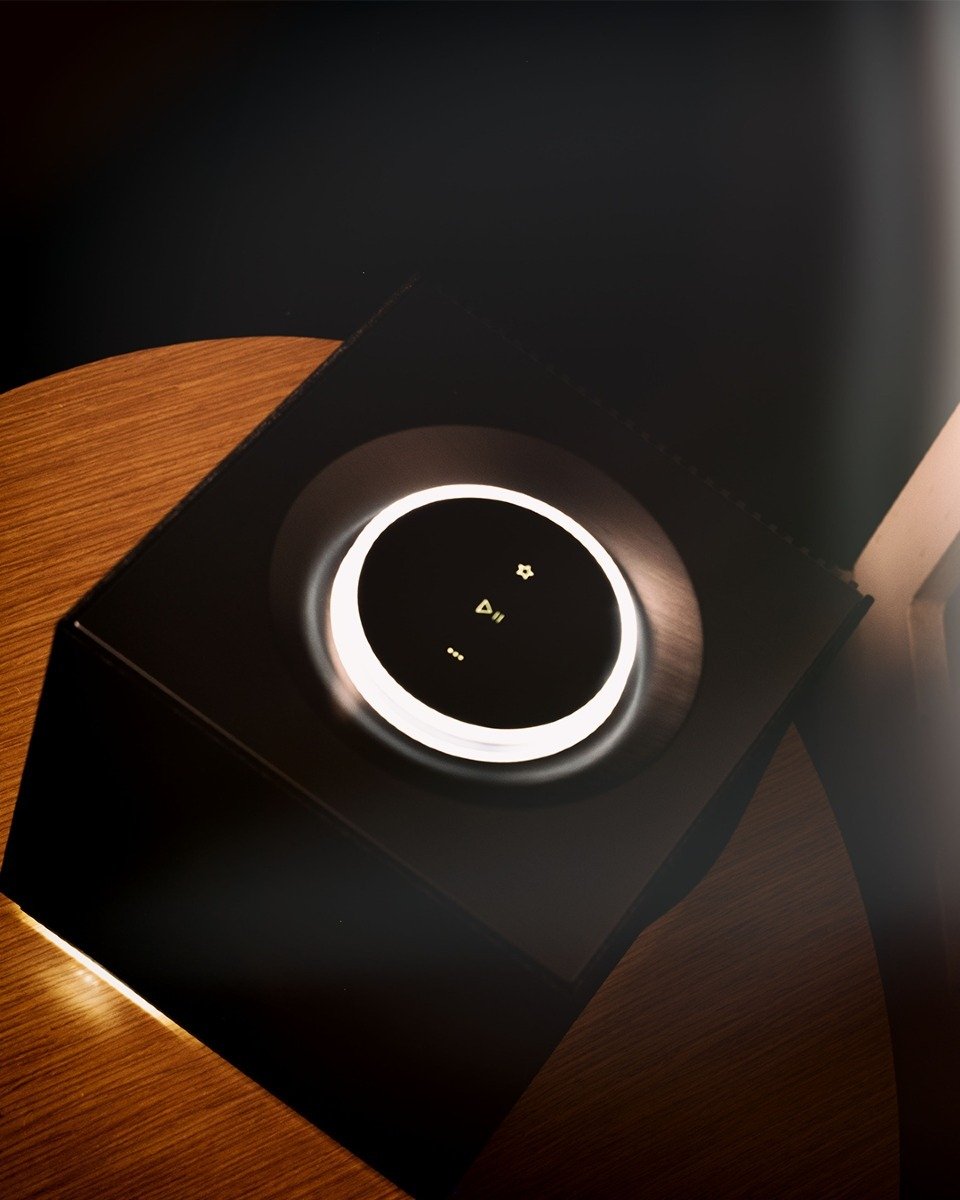Black box with a circular illuminated control dial on a wooden surface, displaying play, star, and ellipsis icons.