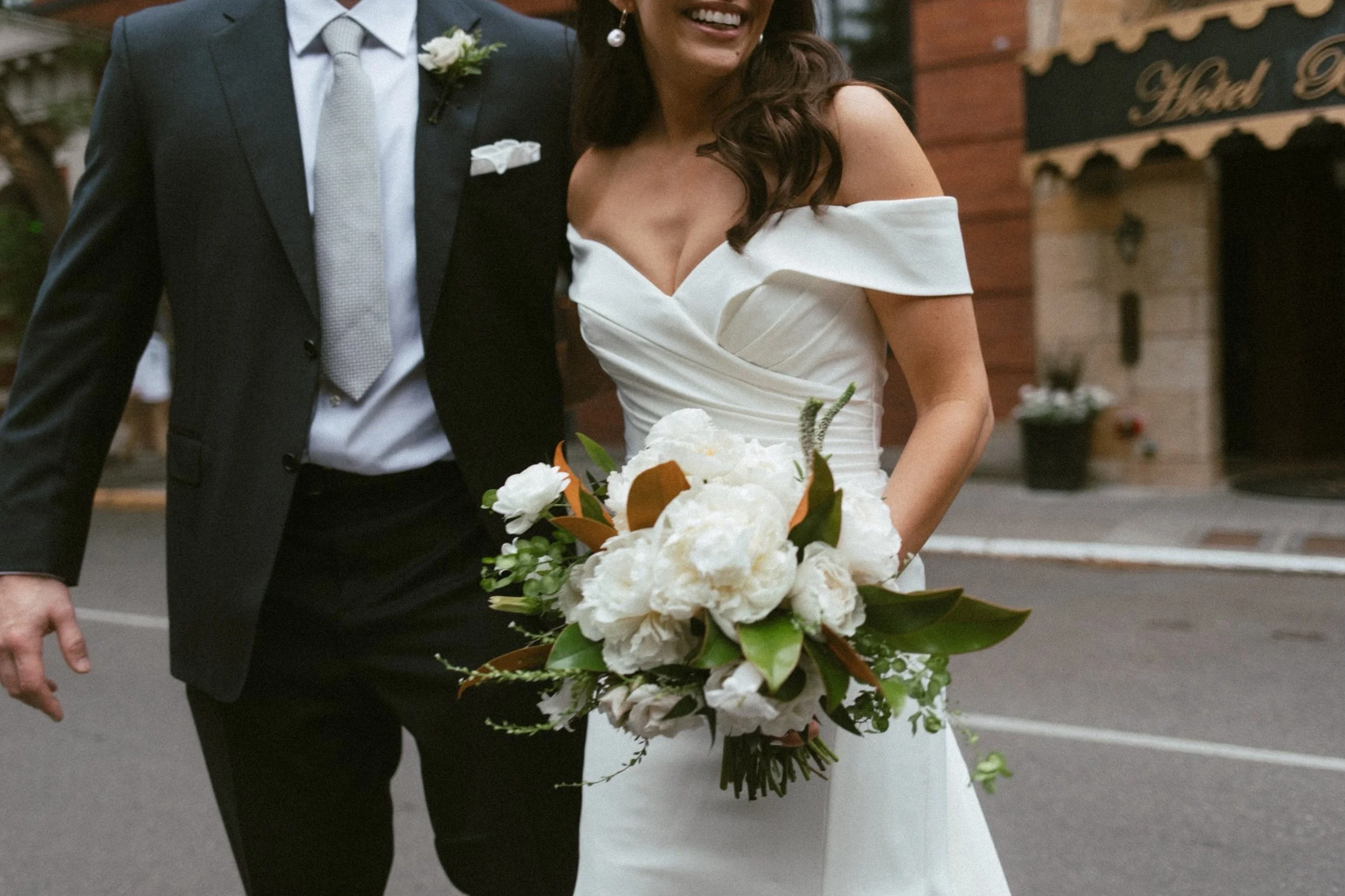 A newlywed bride holding a bouquet of white flowers and wearing an off-the-shoulder white wedding dress, walking beside a man in a dark suit with a light-colored tie. They are smiling and appear to be at a wedding or celebration event.