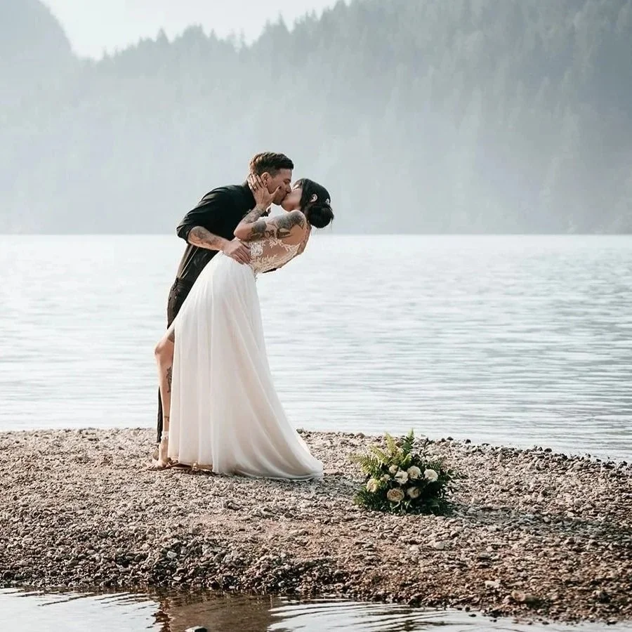 A bride and groom sharing a kiss on a lakeshore, with a bouquet on the ground and forested hills in the background.