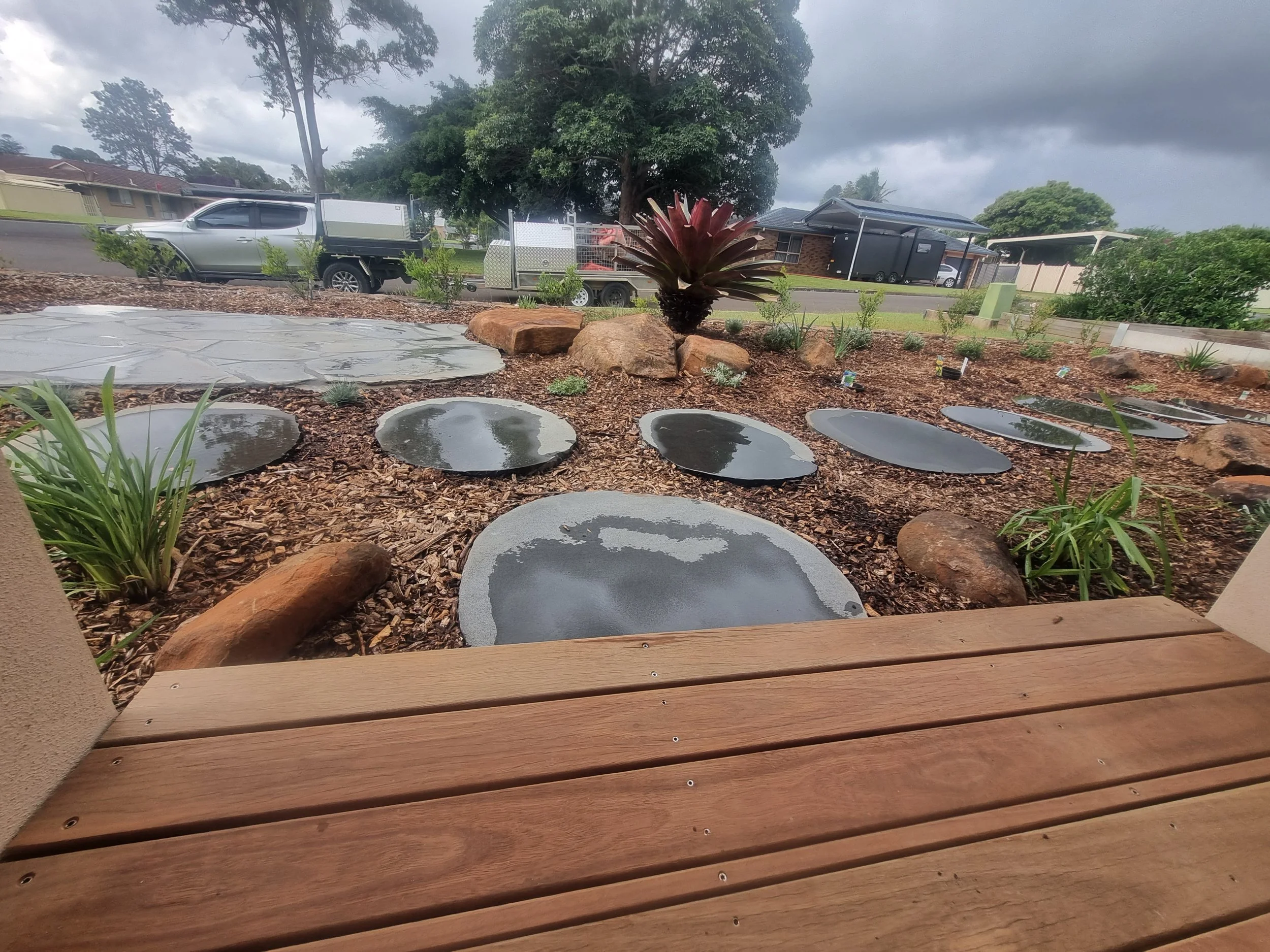 Rainy day view of a landscaped garden with circular stepping stones, a succulent plant, and small bushes, with a wet wooden deck in the foreground and residential houses and a street in the background.