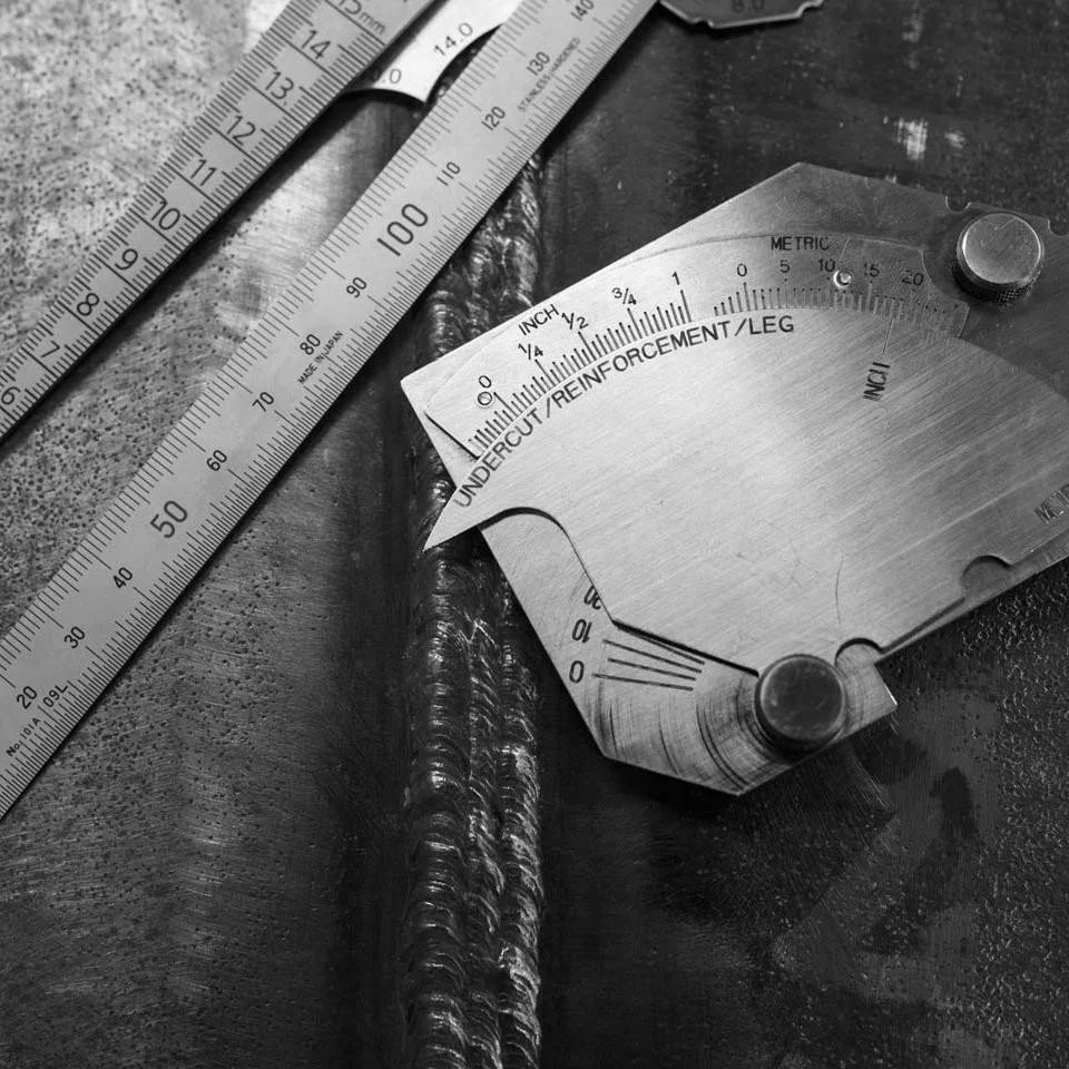 Close-up of a metal measuring tool and a ruler on a dark textured surface, with welding marks visible.