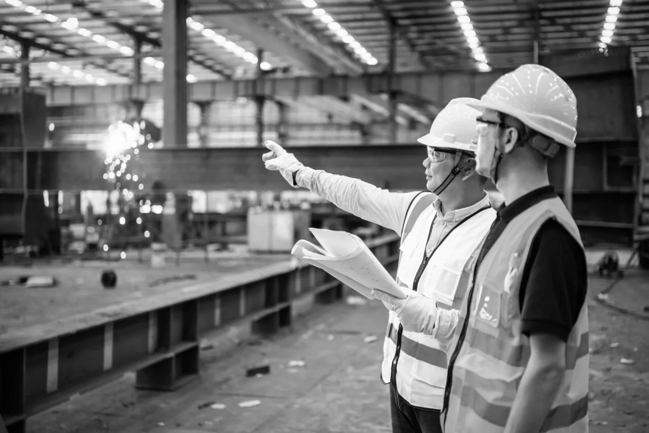 Two construction workers in safety helmets and vests inspecting a construction site, with one pointing and the other holding papers.
