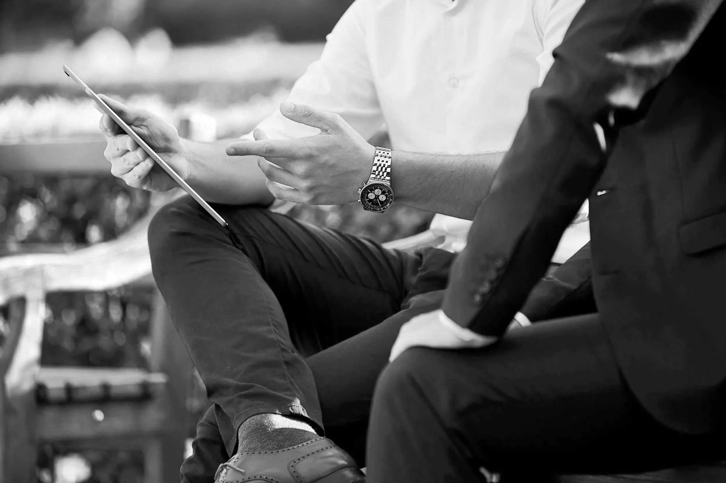 Two men sitting outdoors, one in a white shirt and the other in a dark suit, engaged in conversation. The man in the white shirt is holding a tablet and pointing at it with his fingers. The man in the dark suit is sitting with his hands on his lap.