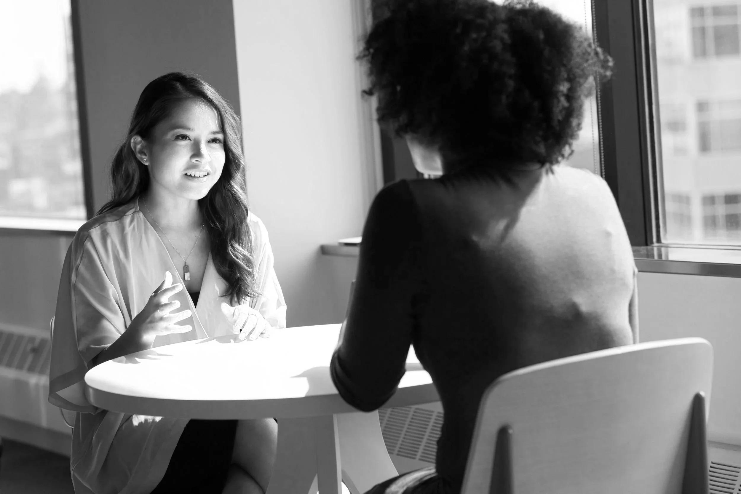 A woman with long wavy hair talking to another woman with curly hair, sitting at a table near a window in a modern office.