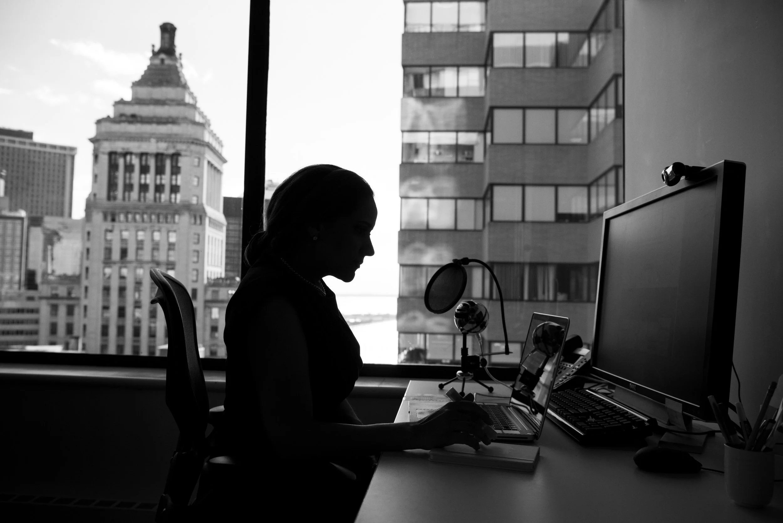 Silhouette of a woman working at her desk in an office with large windows showing city buildings outside.