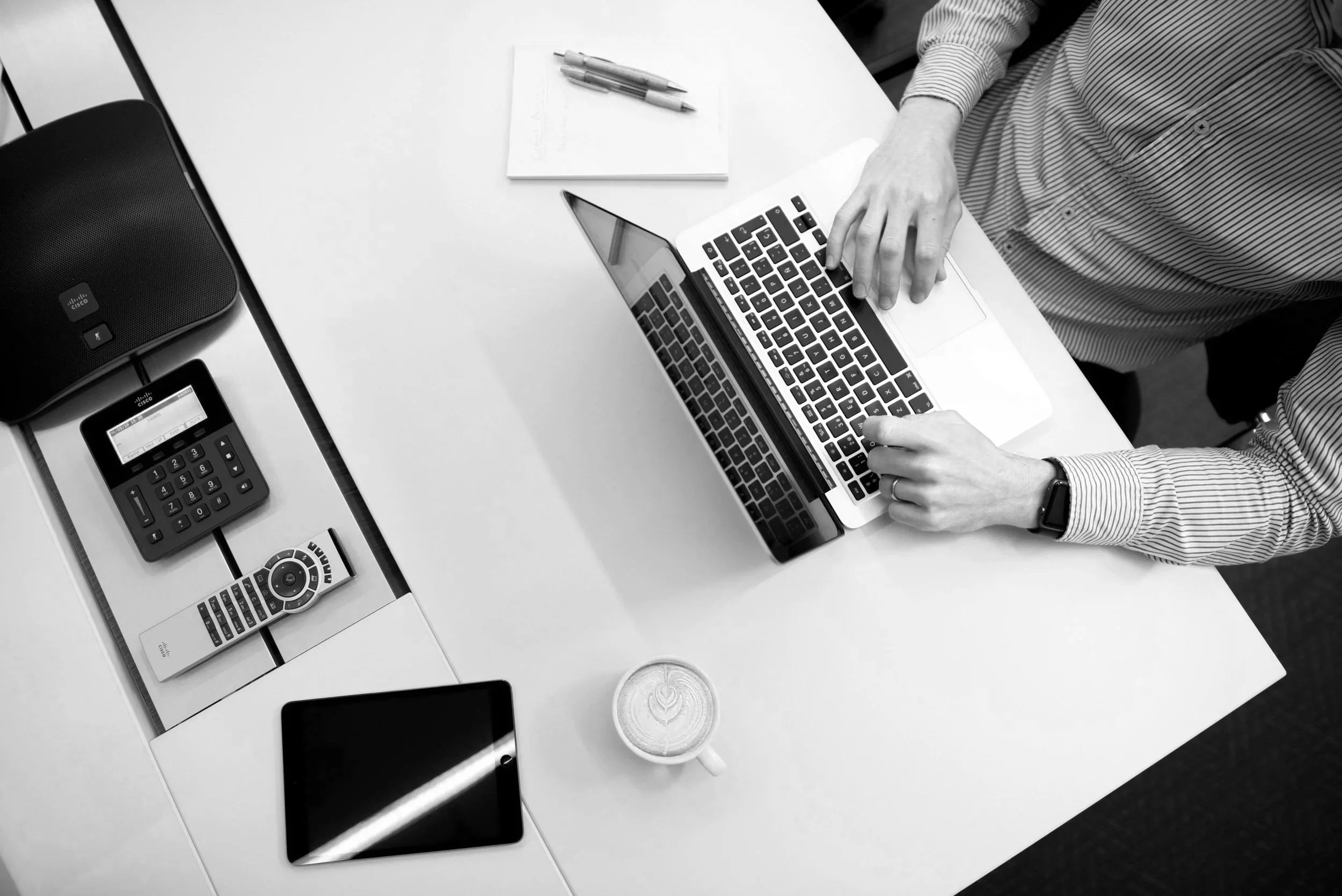 Overhead view of a person working on a laptop at a white desk, with a cup of coffee, smartphone, TV remote, calculator, conference phone, and notepad nearby.