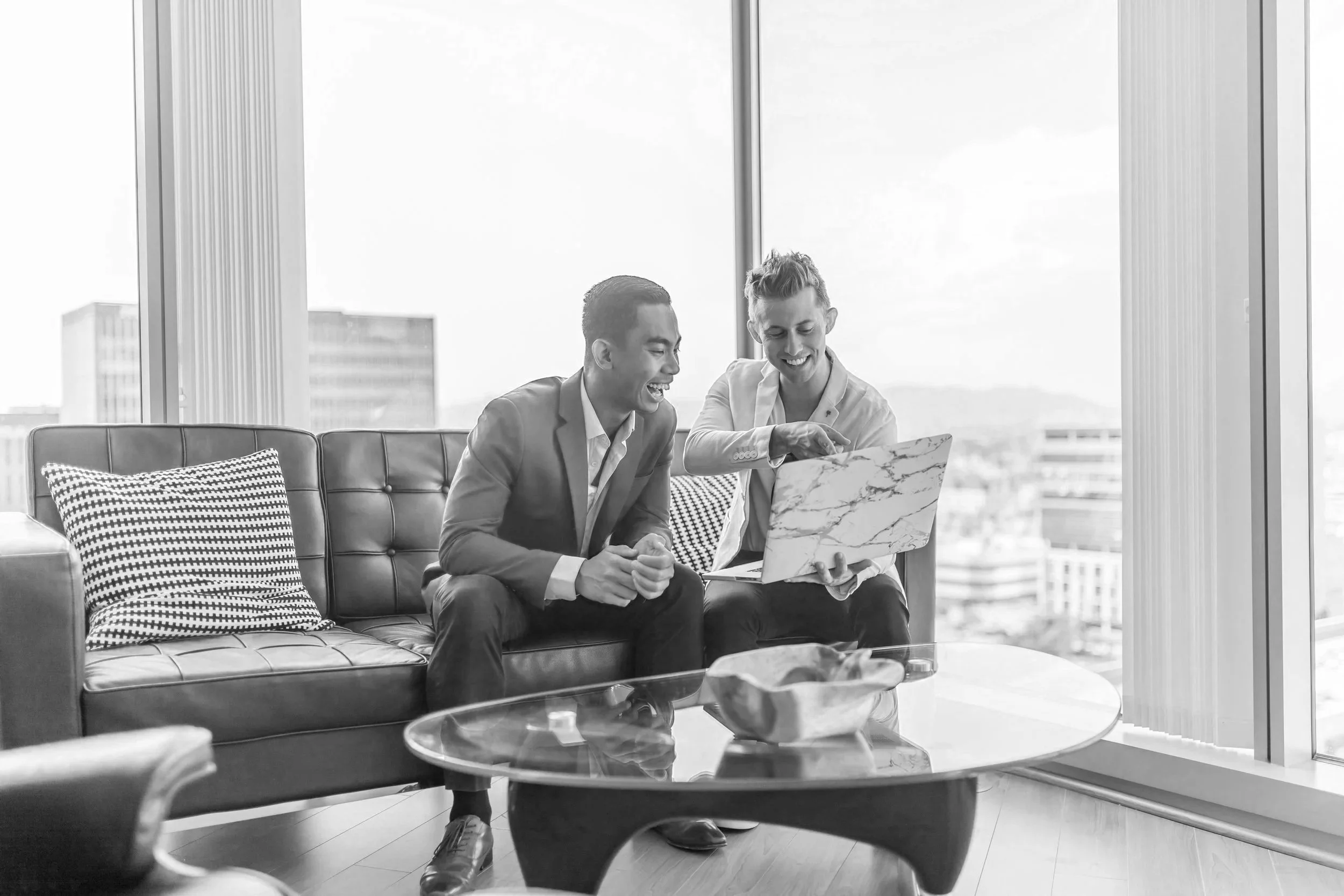 Two men in business attire sitting on a sofa, looking at a laptop, and smiling in a modern office with large windows showing a cityscape.